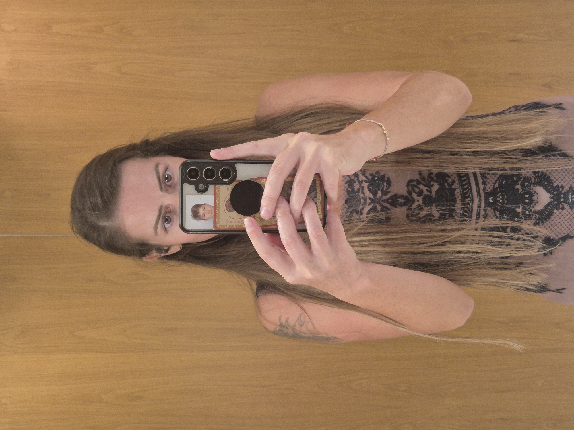 The image displays a woman taking a mirror selfie, with the photograph originally oriented 90 degrees clockwise. When viewed upright, the woman is seen from the chest up, holding a dark-colored smartphone with both hands to capture her reflection.
She has long, straight dark brown hair with lighter highlights, which cascades over her shoulders and upper body. She is wearing a light, possibly beige or muted pink, top adorned with an intricate black lace or patterned overlay. A thin, delicate bracelet with small golden accents is visible on her right wrist, and a subtle, dark-colored tattoo can be seen on her left upper arm. Her brown eyes are focused intently towards the phone's camera, displaying a neutral and concentrated expression.
The smartphone she holds is dark-colored, featuring multiple rear camera lenses and a black circular pop-socket or grip. On the reflective surface of the phone's screen or case, beneath the camera area, there is a small image of a young male's face with short hair. Below this small picture, some unreadable, faint text and numbers are vaguely discernible, possibly part of an identification card or similar item.
The background reflecting the woman is a smooth, light-to-medium brown wooden surface, likely a wall or large paneling. The wood grain patterns run horizontally across the entire frame. The lighting appears soft and even, suggesting an indoor setting during an indeterminate time of day.