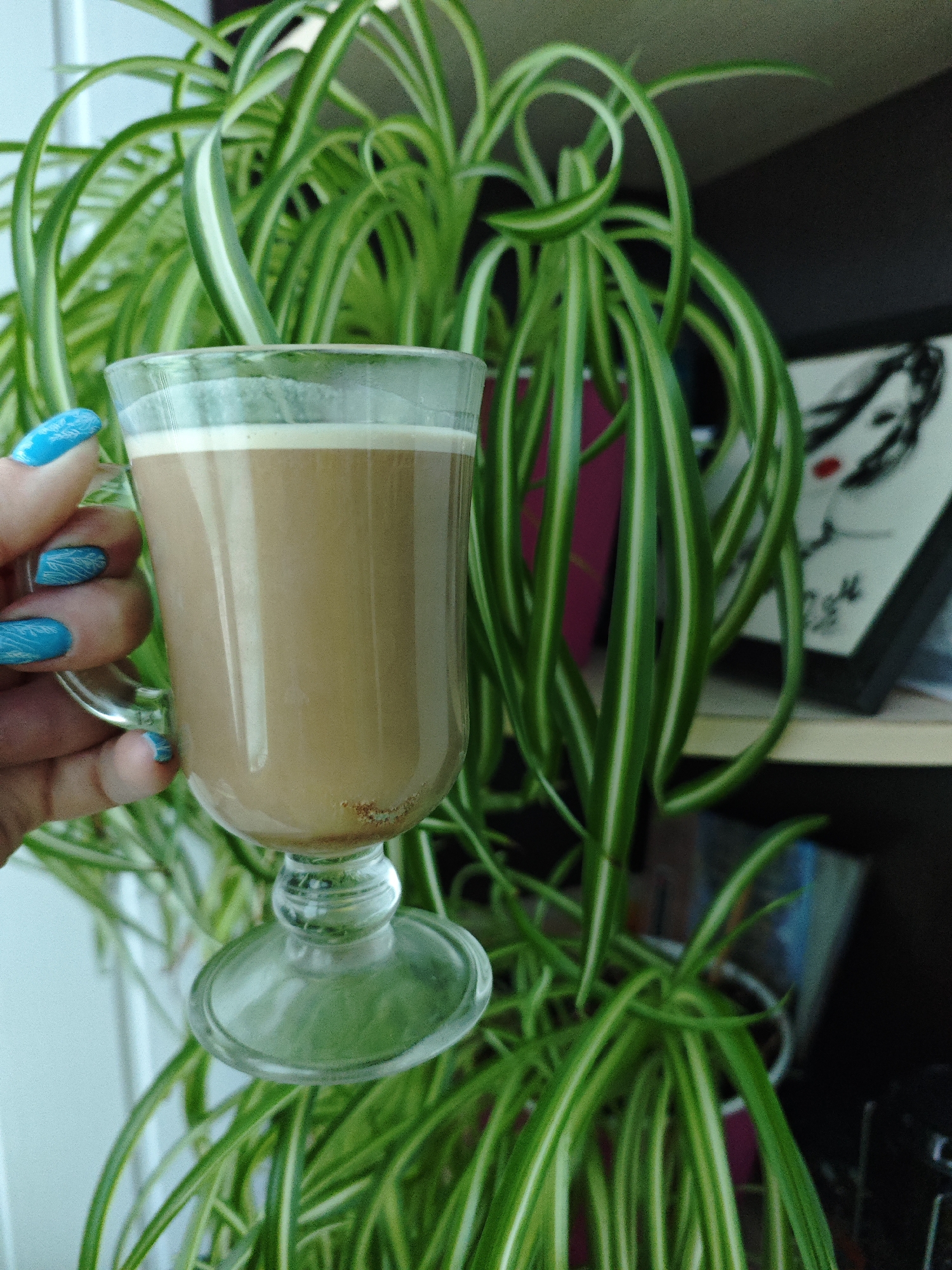 A woman holds a glass mug containing a brown beverage with a thin layer of white foam, possibly coffee or hot chocolate. The woman has bright blue fingernails with a textured, almost scale-like design. The mug is clear glass with a pedestal base. Behind the mug is a lush spider plant with long, curling green leaves featuring white stripes. A shelf is visible behind the plant, containing books and a framed picture with a black-and-white sketch of a woman's face. The location, Markovskii, Russia, cannot be independently verified based on the image content alone.