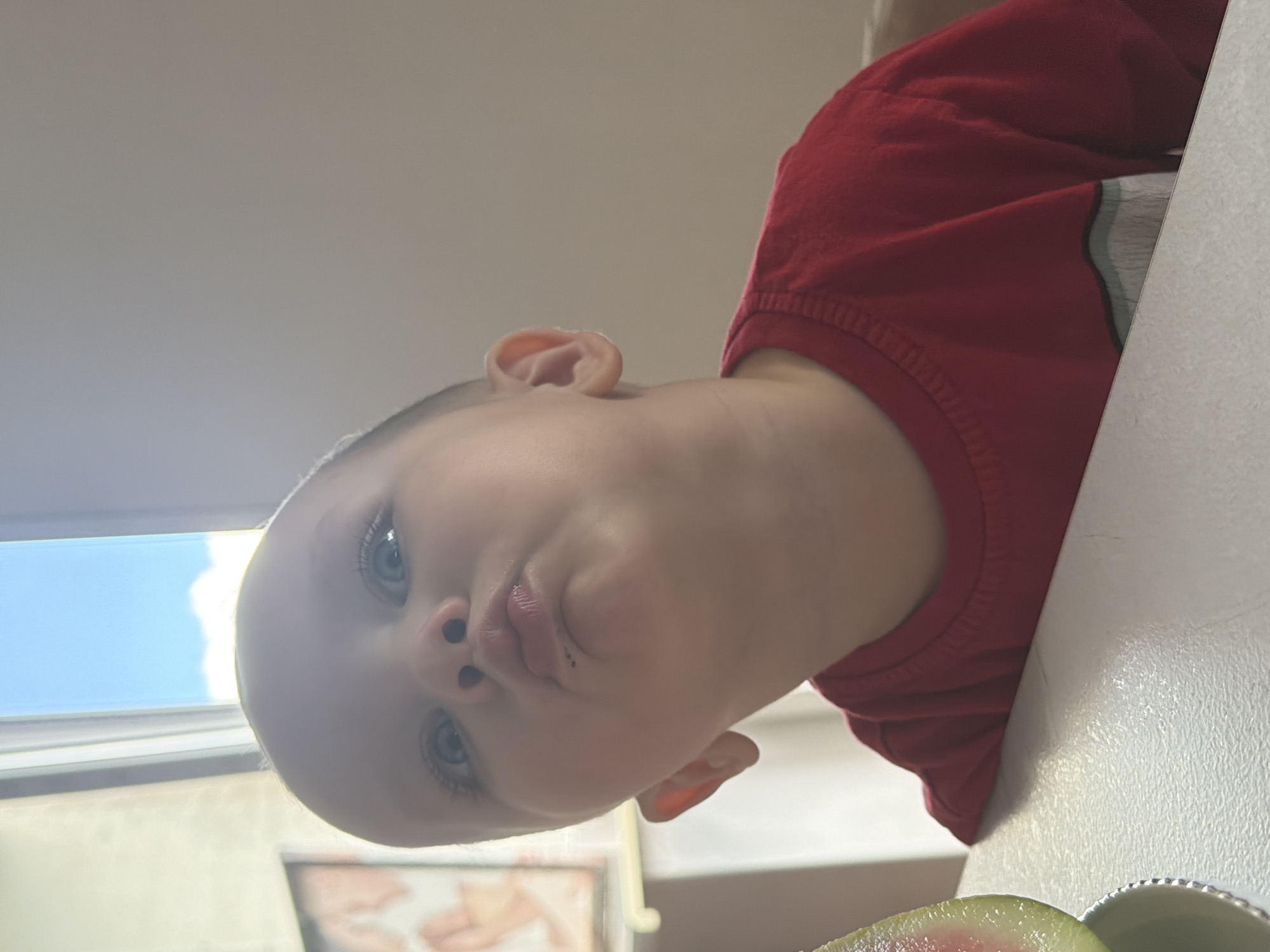 The image captures a young child, appearing to be a toddler, with fair skin and prominent blue eyes, making a puckered facial expression, often referred to as a "fish face." Small dark particles, likely food residue, are visible on the child's lower lip. The child's left ear is visible, and the hair on the sides of the head appears short or closely cropped. The child is wearing a long-sleeved, crew-neck red shirt, with a horizontal light-colored stripe visible near the bottom edge.
In the immediate foreground on the left, a slice of watermelon, showing its red flesh and green rind, rests in a light-colored bowl with a distinct, possibly beaded, rim. The child is seated at a white, textured surface, likely a table or high-chair tray.
The background reveals an indoor setting with natural light. To the left, a large window offers a view of a clear blue sky with some scattered white clouds. Below the window, part of a wall and a framed piece of artwork or photograph, displaying abstract shapes or possibly stylized human forms in warm hues, is partially visible. The background to the right of the child consists of a plain, light-toned wall. The scene is described as being located in the city of Chaikovsky, Russia.
