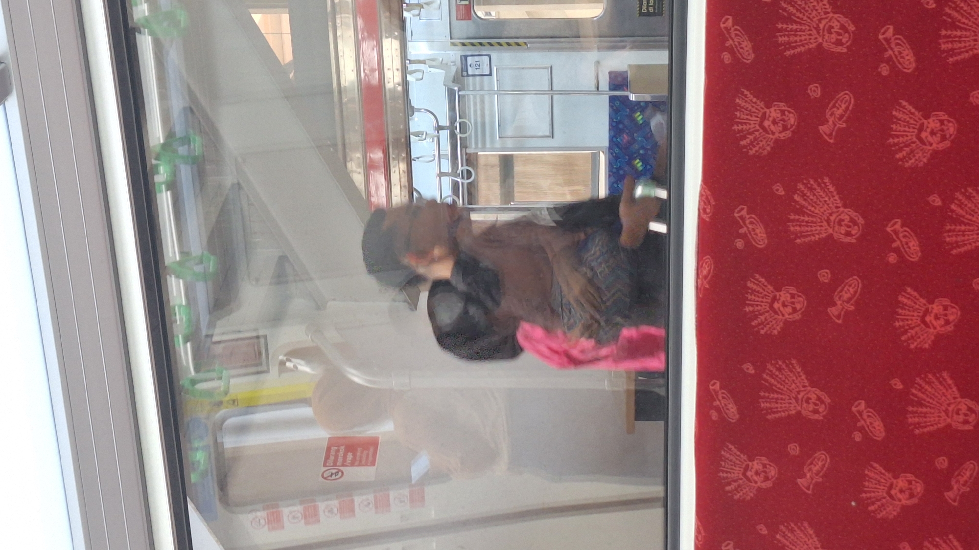 The image depicts the interior of a commuter train, likely in Cikarang, Indonesia, given the context. The scene is viewed through a window, showing reflections that add to the visual complexity.
The primary subject is a person, likely a woman, seated inside the train. She has long, brown hair and is wearing glasses and a dark jacket over a patterned shirt. Her body is mostly obscured, but she appears to be holding a pink cloth or garment in her right hand. Her facial expression is not clearly discernible due to the angle and reflections, but her posture suggests she might be resting or waiting.
The train car interior features typical commuter train elements: handrails, seats covered in red fabric with a repeating pattern of what appear to be stylized heads and musical instruments, and windows. Some of these handrails are green and appear to be made of plastic. Reflections in the window show other passengers or elements of the train's exterior, and the bright lighting suggests it is daytime.
Visible text includes a red sign with white text and graphics on the left side of the frame, partially obscured by reflections. One snippet of text that is legible on a sign inside the train car is "121". Another red sign with white lettering reads "Dilarang Merokok" which translates to "No Smoking" in Indonesian.
The overall impression is a mundane moment captured during a train journey, with the reflections creating a somewhat abstract and layered visual experience. The bright red seating adds a vibrant color contrast to the otherwise metallic and grey tones of the train's interior.
