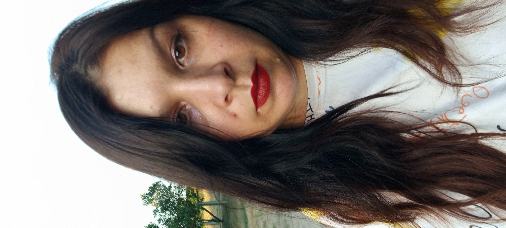 A close-up, front-facing, eye-level selfie of a woman with long, dark brown hair and brown eyes. She is wearing red lipstick and has noticeable makeup, including shimmering eyeshadow. Her skin appears to have freckles and some blemishes. She is wearing a white t-shirt with colorful markings on it. The background is slightly blurred, showing trees, a fence, and a grassy area, suggesting an outdoor, possibly rural or suburban, setting. The lighting indicates it is daytime. There is no specific identifiable information to place this image in Klyntsi, Ukraine.
