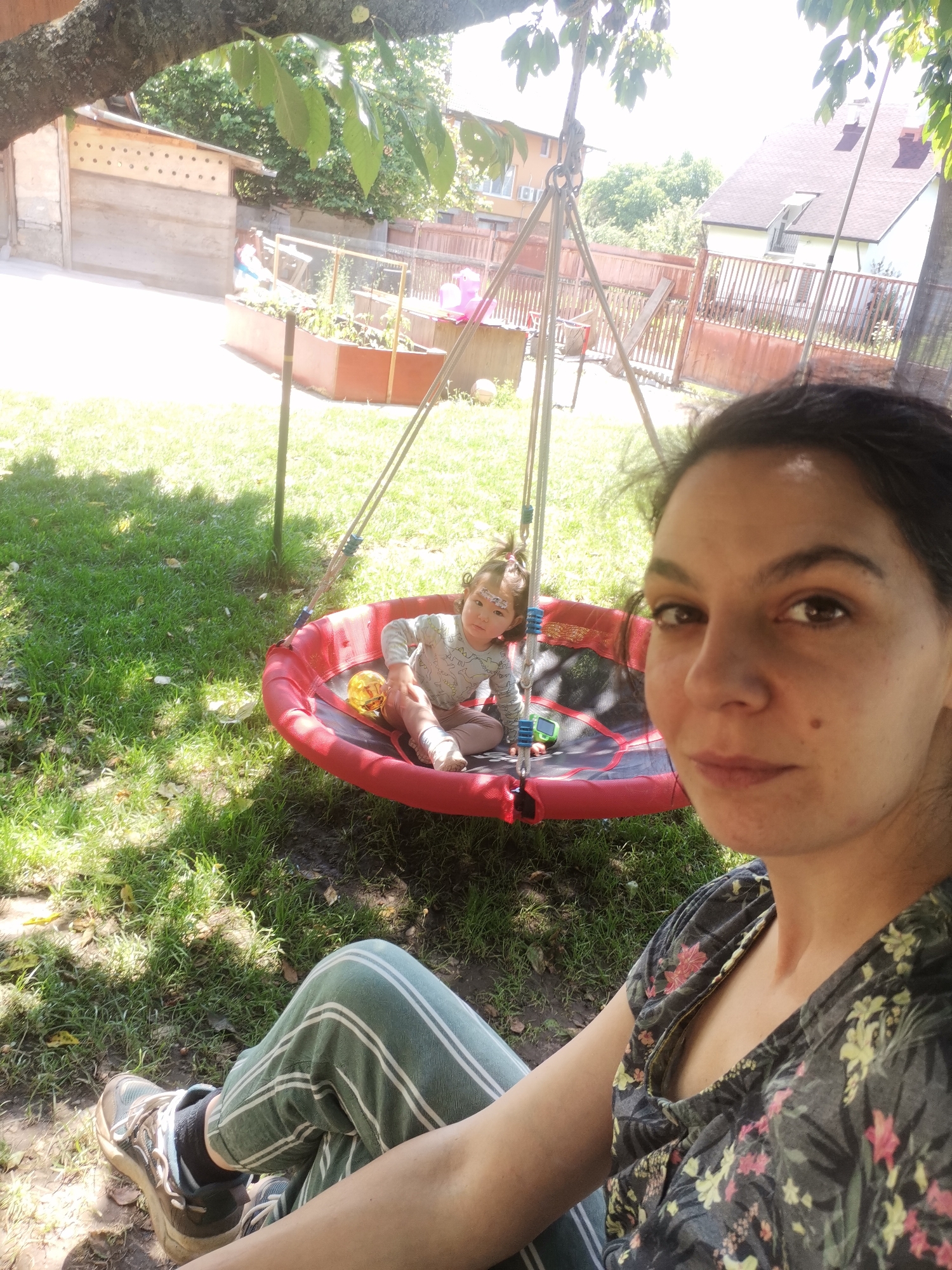 A medium shot shows two people outdoors on a sunny day, with one person in the foreground and another further back in a circular swing. The person in the foreground is a woman with dark hair and brown eyes, looking at the camera. She is wearing a dark, floral-print shirt and green and white striped pants, with a sneaker on her left foot. The person in the background is a young child with dark hair, sitting on the swing. The child is wearing a light-colored long-sleeved shirt and light-colored pants, and has some face paint on their forehead. The swing is red and black, and is suspended from a metal frame. The background shows a grassy yard with trees, a wooden fence, and a building that appears to be a house. The scene appears to be a residential backyard in Osoitsa, Bulgaria.