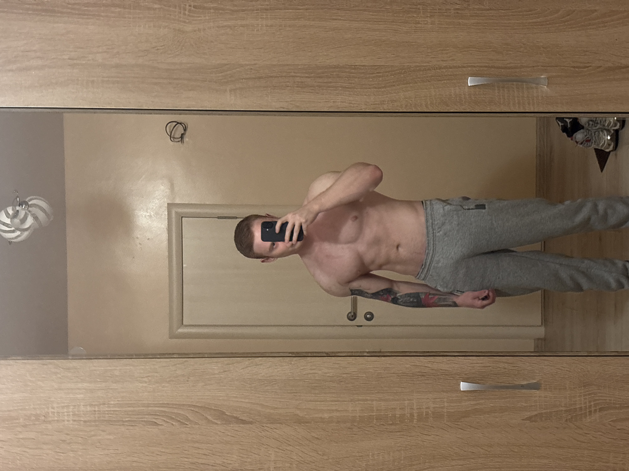 The image shows a shirtless man wearing gray sweatpants taking a selfie in a mirror. The mirror is framed by a wooden dresser. In the background, there is a door. The environment appears to be a bedroom in a house or apartment. It's impossible to determine the exact location in Chaikovsky, Russia from the information provided.