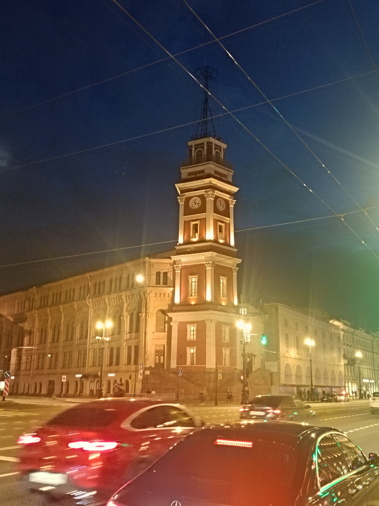 The image captures a night scene in Saint Petersburg, Russia, featuring the City Duma Tower, a distinctive architectural landmark with a clock tower. The tower is illuminated and stands against a dark blue sky. In the foreground, blurred cars move along a street, suggesting traffic. Streetlights add to the urban ambiance. The building adjacent to the tower has many windows and is also illuminated. Overhead wires stretch across the sky. There are a few people on the sidewalk near the base of the tower.