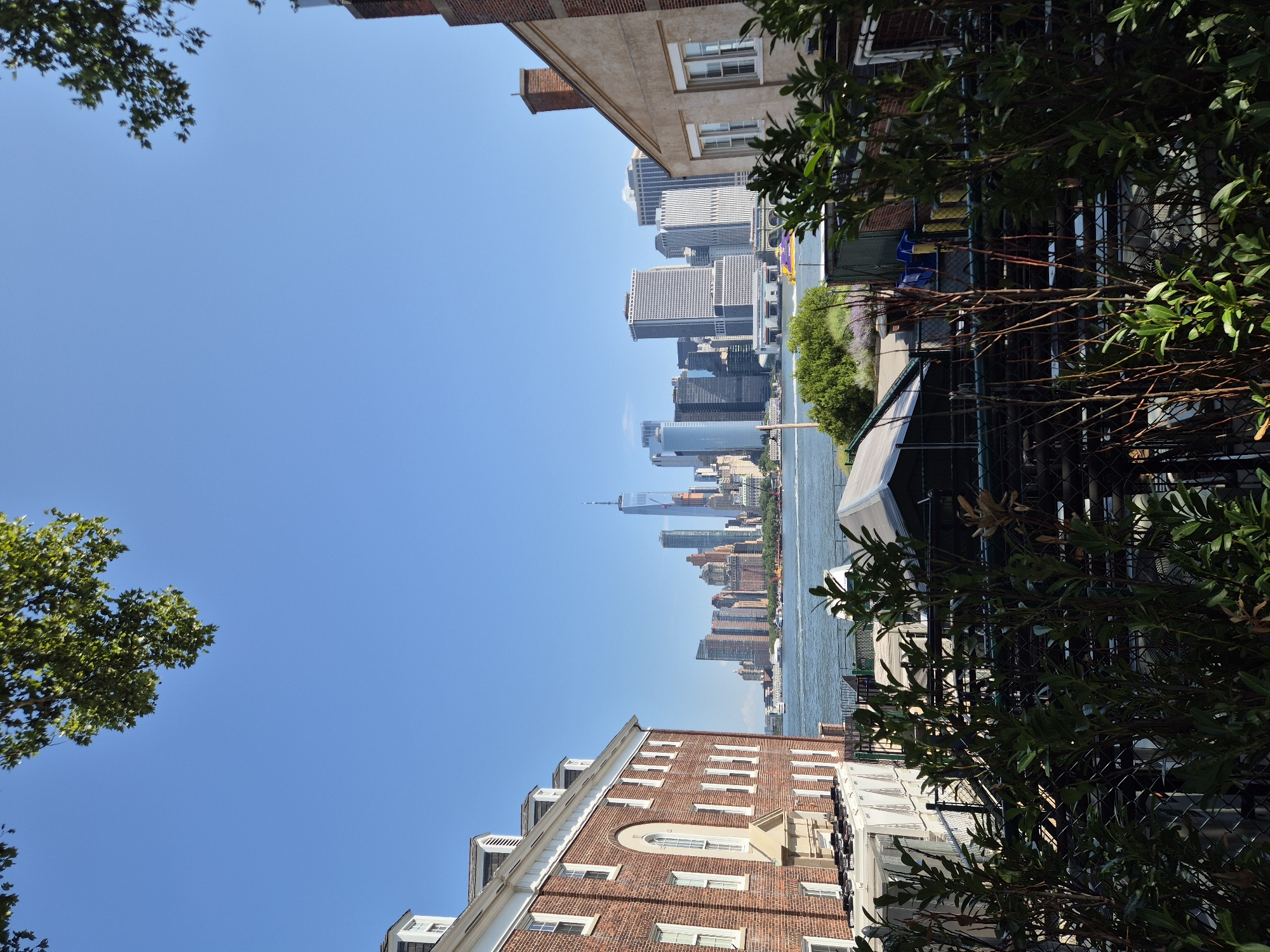 The image depicts a view of the Lower Manhattan skyline from across the water, likely from Governors Island, New York City. In the foreground, on the left, is a red brick building with multiple windows and dormers. To the right, a portion of a tan stucco building with a brick chimney is visible. Between these buildings, a body of water, the New York Harbor, separates the foreground from the distant cityscape. The skyline is dominated by numerous skyscrapers of varying heights and architectural styles, including the iconic One World Trade Center. A bright yellow and purple ferry or boat is visible on the water. Lush green trees and foliage partially obscure the lower portion of the scene. The sky is a clear, bright blue, indicating a sunny day.