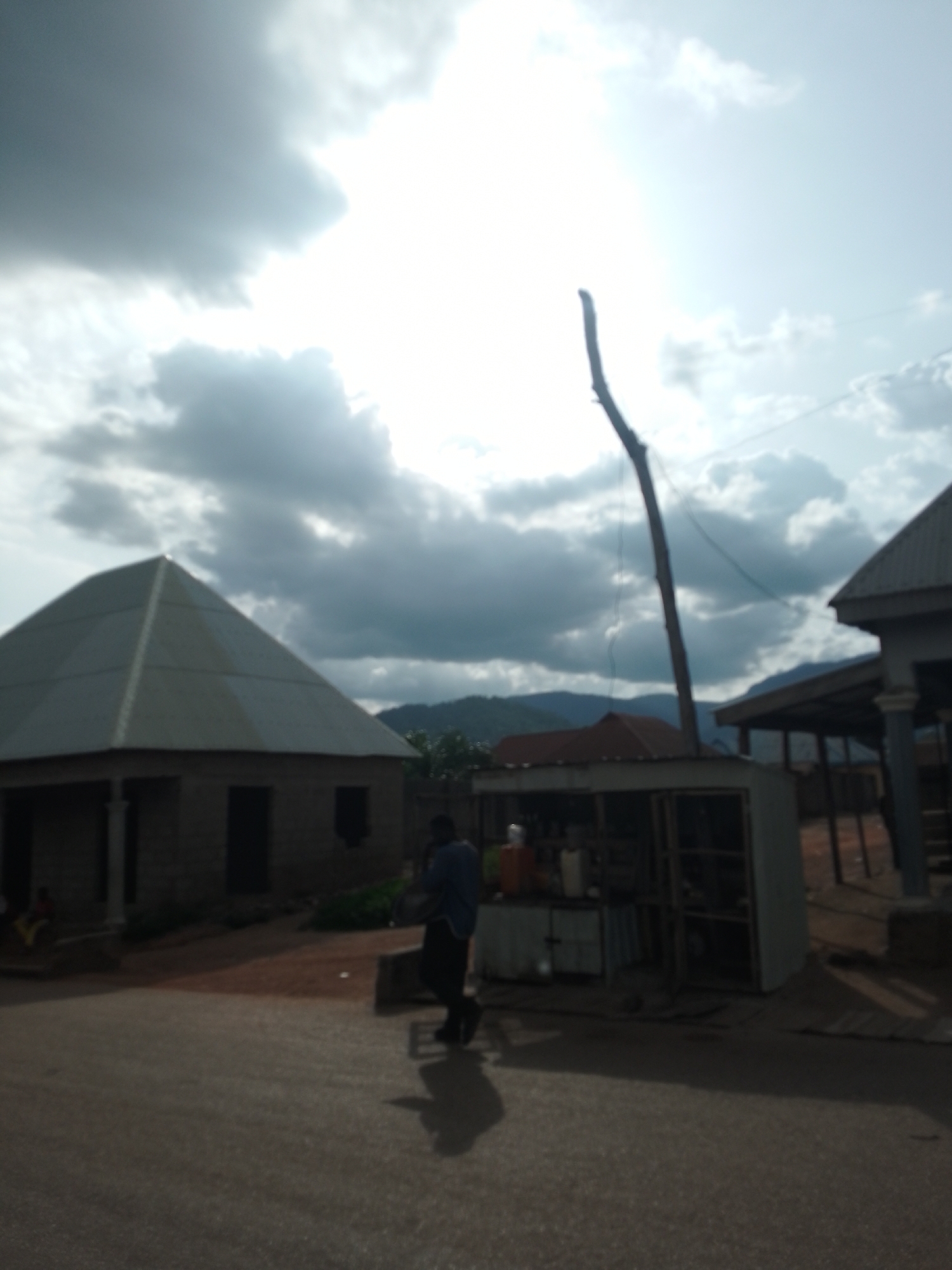 The image depicts a street scene in Serti, Nigeria, under a cloudy sky with bright sunlight filtering through. In the foreground, a paved road is visible, with shadows cast from structures and a person. A person wearing a dark blue shirt and black pants stands on the road, facing away from the camera and towards a small roadside stall. The stall is open-fronted, showcasing various items, possibly goods for sale, including bottles and containers. Behind the stall, a weathered wooden pole with a diagonal brace extends upwards towards the sky, likely part of the stall's structure or a utility pole. To the left of the stall, a hexagonal-roofed building with a dark entrance is prominent, its structure appearing to be made of brick or concrete. Further in the background, other buildings and structures are partially visible, suggesting a residential or commercial area. Distant hills or mountains are faintly discernible against the bright sky, which is dominated by dramatic clouds and intense sunlight. Another person is seated on the ground near the hexagonal building, a smaller figure in the distance.