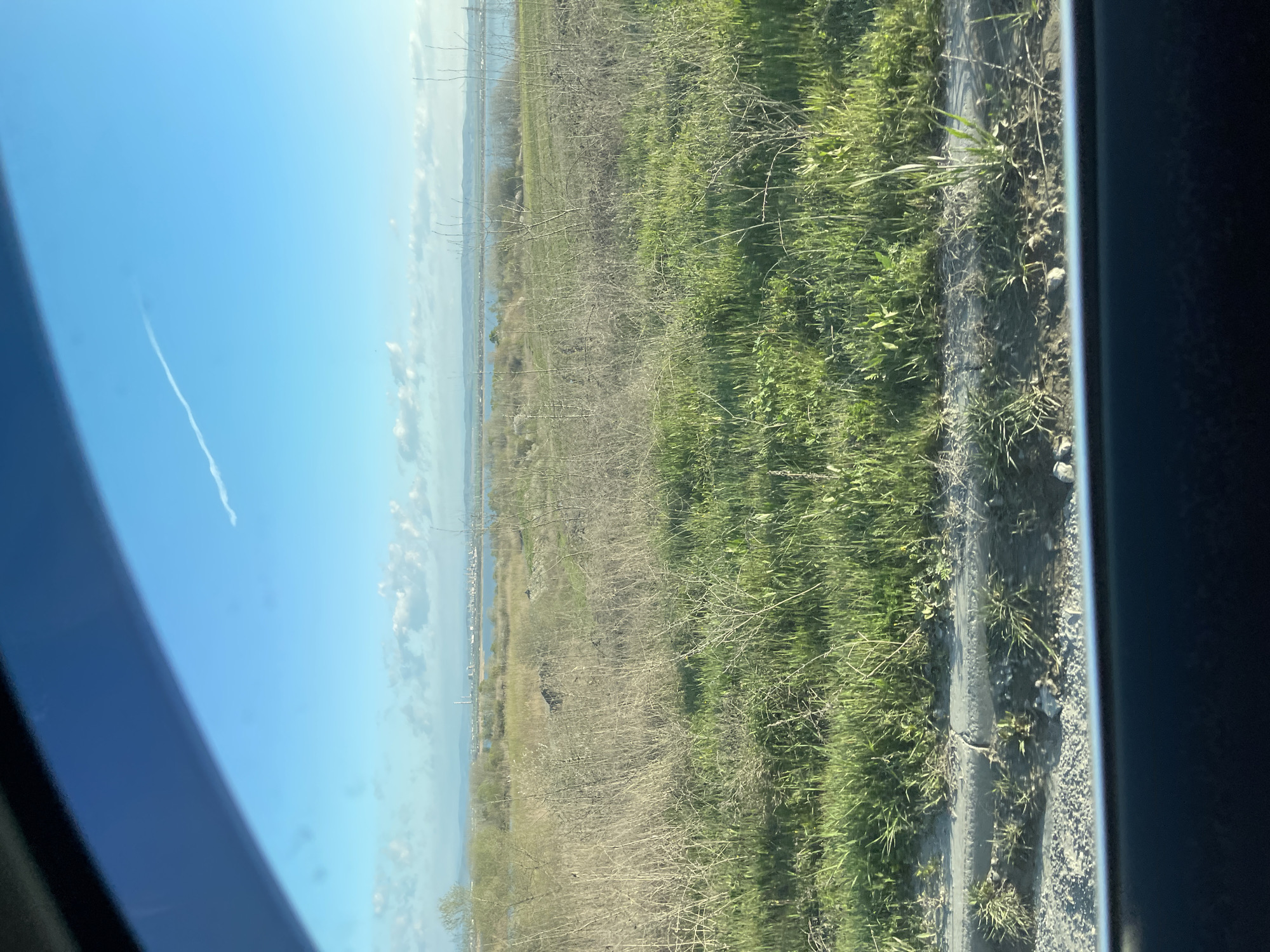 The image shows a view from inside a vehicle, looking out over a landscape in Burgas, Bulgaria. The sky is blue with a contrail of a plane visible. A body of water, potentially part of the Burgas lake system, is visible in the distance. The foreground consists of grassland vegetation and roadside terrain. A dark object, possibly an animal or rock, is visible in the mid-ground vegetation. The window frame of the vehicle is seen along the left and right sides of the image.