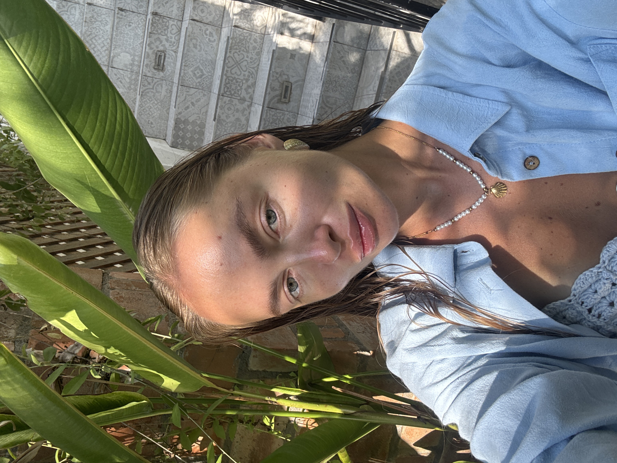 The image is a selfie of a woman in Unknown, Indonesia. She is wearing a light blue collared shirt and a pearl necklace with a seashell pendant. Her hair is wet. The background consists of green tropical plants and a brick wall. In the upper background, there are stairs with patterned tiles. The lighting is bright and sunny, casting shadows on the subjects.
