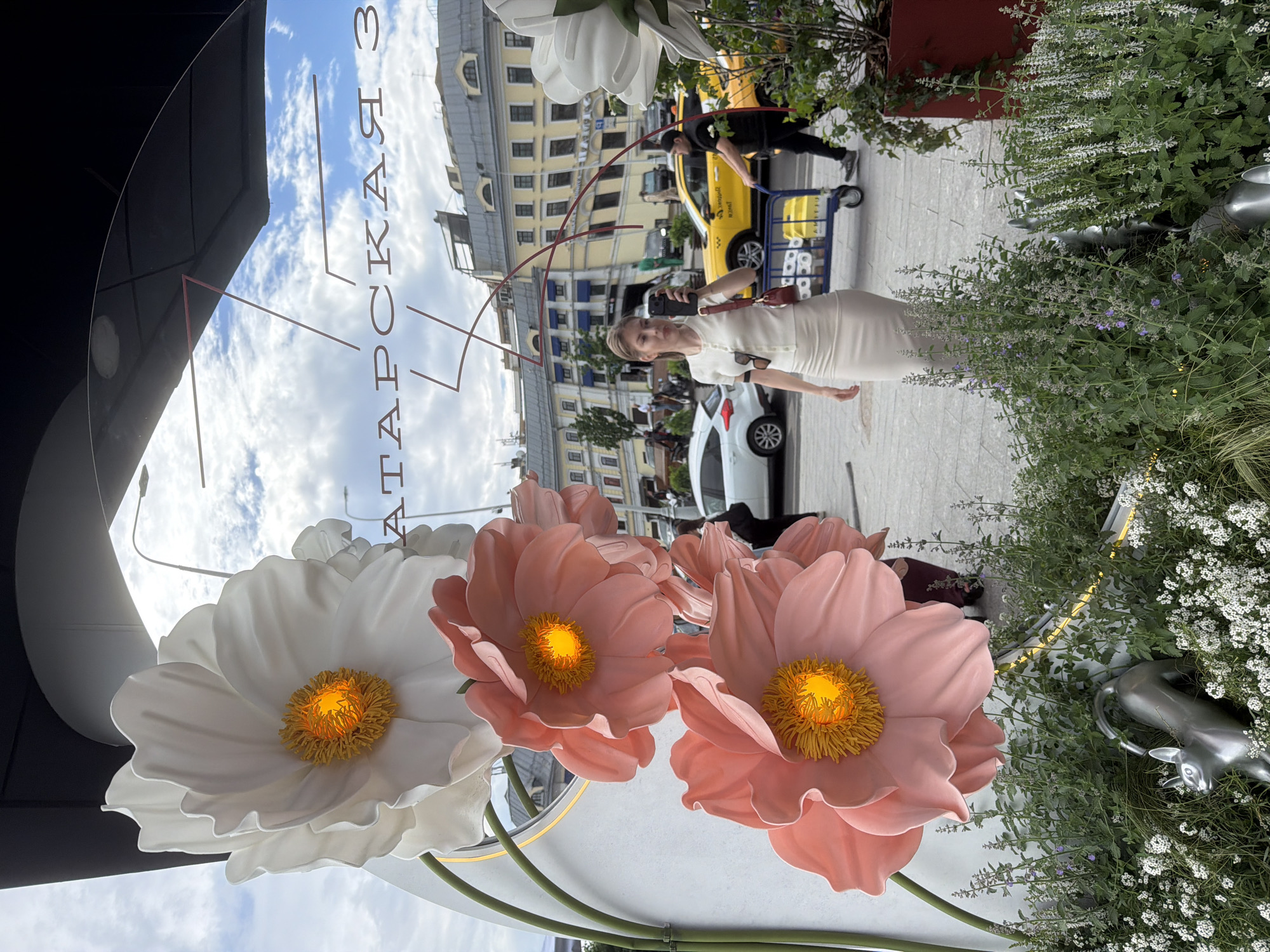 The image, taken in Moscow, Russia, features a woman in a white dress holding a black phone, apparently taking a picture of herself. She is standing on a sidewalk near a building with a storefront display featuring oversized white and pink flowers. Behind her, a yellow taxi is visible, along with a man pushing a blue hand truck, and a white car. The environment includes greenery with small white flowers. Reflected on the store's sign is the sky with fluffy white clouds. The street sign reads "Tatarskaya 3".