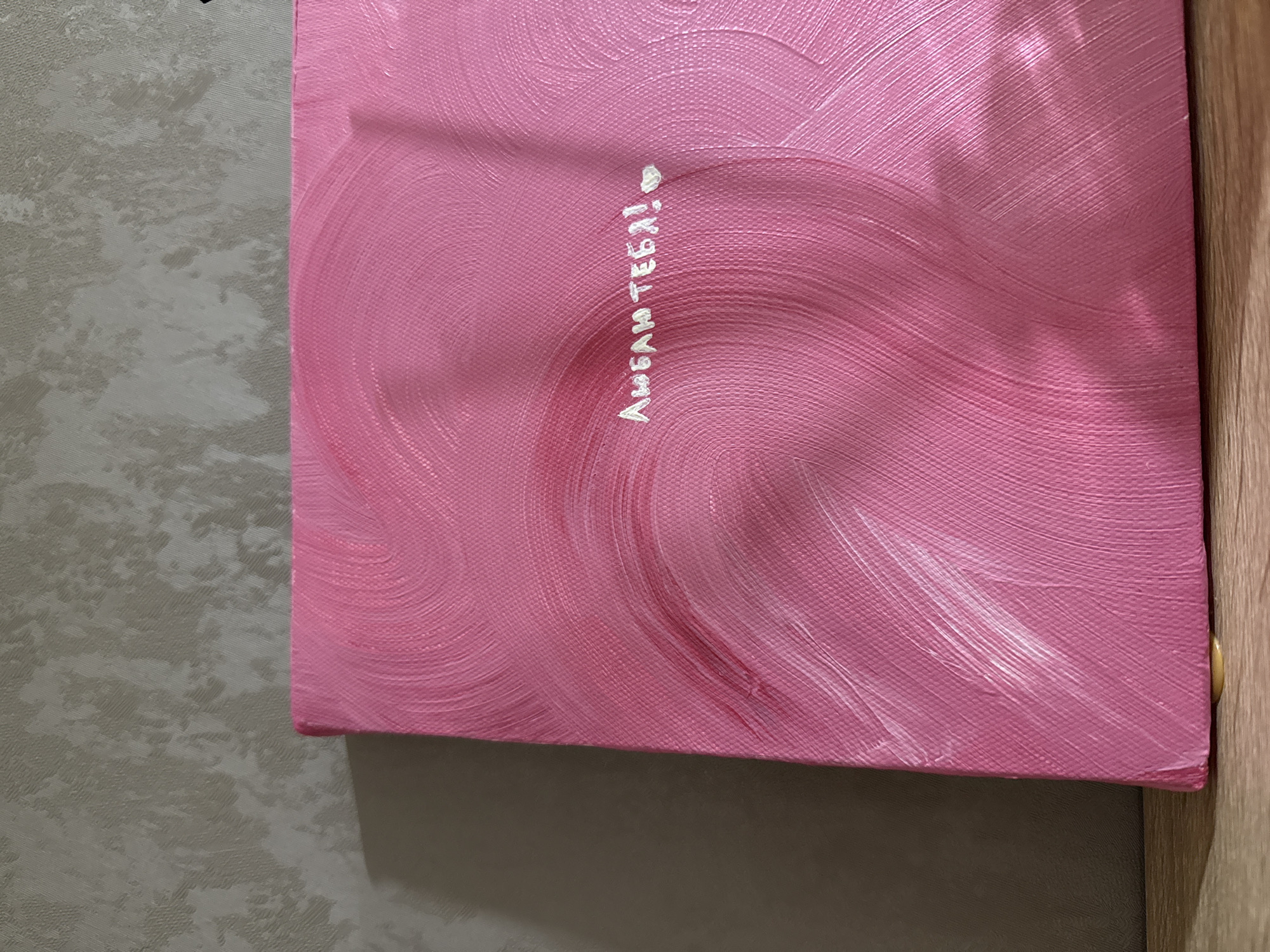 The image shows a close-up of a pink painted canvas, possibly oil or acrylic, with visible brushstrokes. The canvas is positioned against a wall, and a small portion of a wooden structure is visible on the right edge. Written vertically on the canvas in white paint is the Russian phrase "Люблю тебя!", which translates to "I love you!". A small heart is painted next to the phrase. The image doesn't provide any visual clues or details to pinpoint the location as Chaikovsky, Russia.