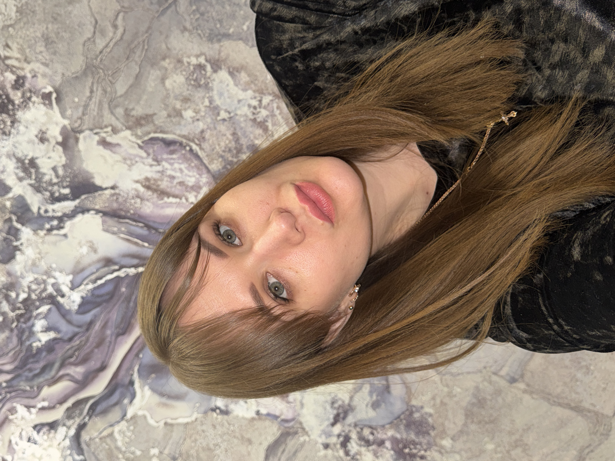 A young woman is featured in a close-up portrait, captured from a slightly overhead and angled perspective, with her head tilted. She has long, straight, light brown or auburn hair that frames her face and cascades down her side. Her eyes are a prominent green, and she looks directly at the camera with a calm expression and a subtle, gentle smile. She wears light makeup, including a soft pink lip color. Visible details include multiple small, shiny earrings in her right ear and a thin gold-colored chain with a small cross pendant resting near her neck, partially intertwined with her hair. She is dressed in a dark, textured garment, possibly made of velvet or crushed velour. The background consists of a patterned surface, likely a decorative floor or wall, characterized by swirling veins of grey, white, and muted purple, resembling marble or natural stone. The photograph appears to be taken indoors in Chaikovsky, Russia.
