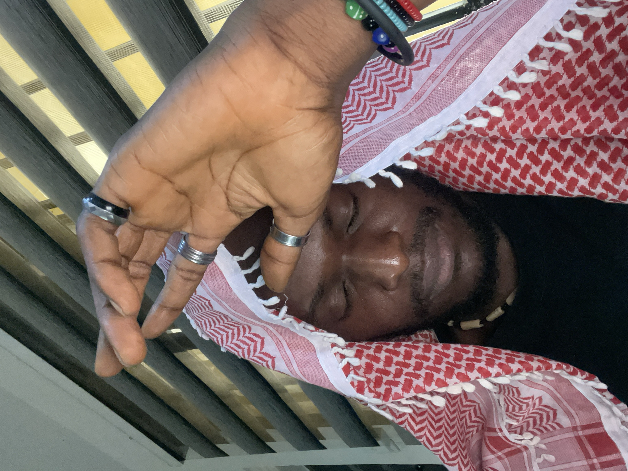 A close-up shot captures a man wearing a red and white checkered keffiyeh. His eyes are closed, and his right hand, adorned with multiple rings and a bracelet, is raised to his forehead. He is wearing a black shirt and a beaded necklace. The background consists of horizontal blinds. The image does not contain any information to suggest a location in Abuja, Nigeria.