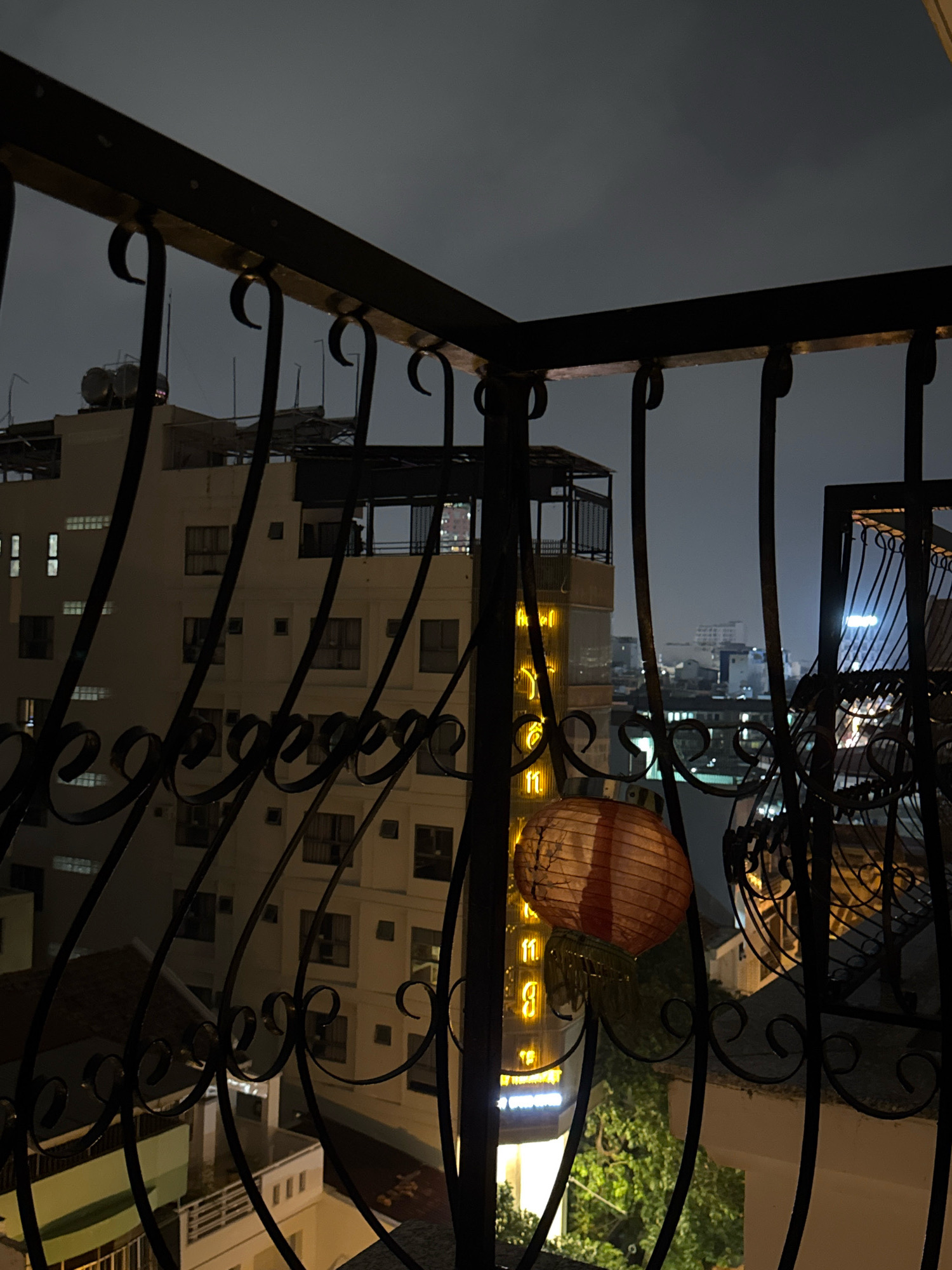 The image depicts a nighttime view from a balcony in Nha Trang, Vietnam, looking out over a city skyline. A dark, ornate metal railing frames the foreground. In the middle ground, a multi-story building with many windows is visible, some illuminated from within. A few other buildings, some with illuminated signage, are scattered across the cityscape. A round, paper lantern, lit from inside, hangs from the railing in the lower left of the frame. The sky is dark and overcast. There are no people visible in the image.