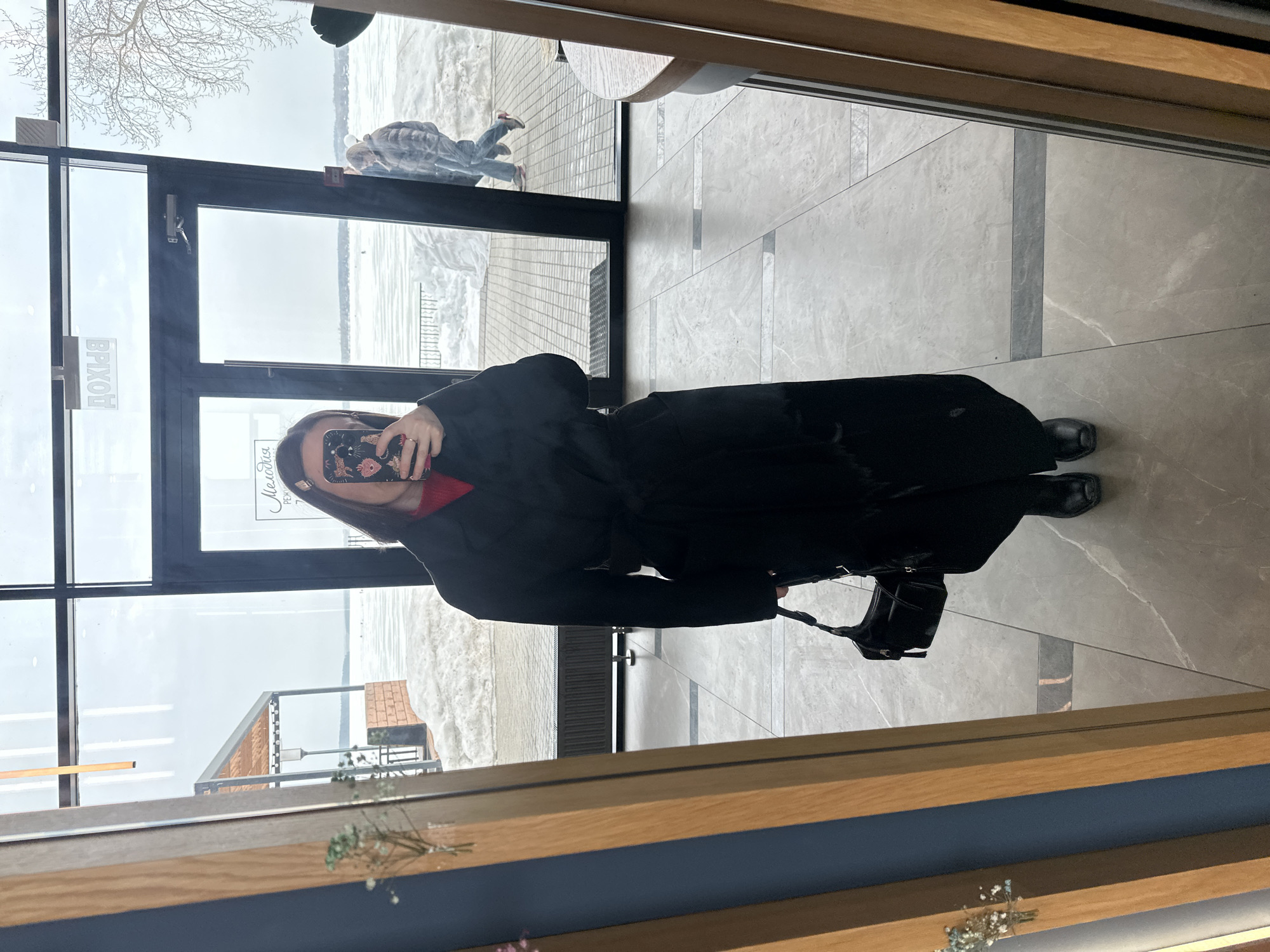 A full-length mirror selfie captures a person standing indoors on a light gray tiled floor with dark gray linear accents. The individual, presumed female, is dressed in a long, belted black coat over a red top, black boots, and holds a black shoulder bag with a tassel. A smartphone with a floral-themed case is held in their right hand, covering their face. A ring is visible on the fourth finger of the left hand. Small white flowers in a vase are reflected in the mirror's left wooden frame.
Through large, black-framed windows, an exterior scene is visible in Votkinsk, Russia. The immediate outdoor area features a paved surface with patches of snow. Beyond this, a wide, snow-covered, likely frozen body of water (Votkinsk Reservoir) extends to a distant shoreline. An outdoor pergola-like structure with a light roof is visible to the left. The sky is overcast. A bare deciduous tree is visible through the upper right window pane. Reflected on the window are partially visible Russian words, including "ДОХОД" (DOKHOD - entrance/income) and "Мелодия" (Melodiya - Melody), suggesting the indoor location is a commercial establishment, possibly a cafe. Two individuals are seen walking outdoors on the paved area, partially obscured by the window frame; one wears a light puffer jacket, blue jeans, and pink shoes.