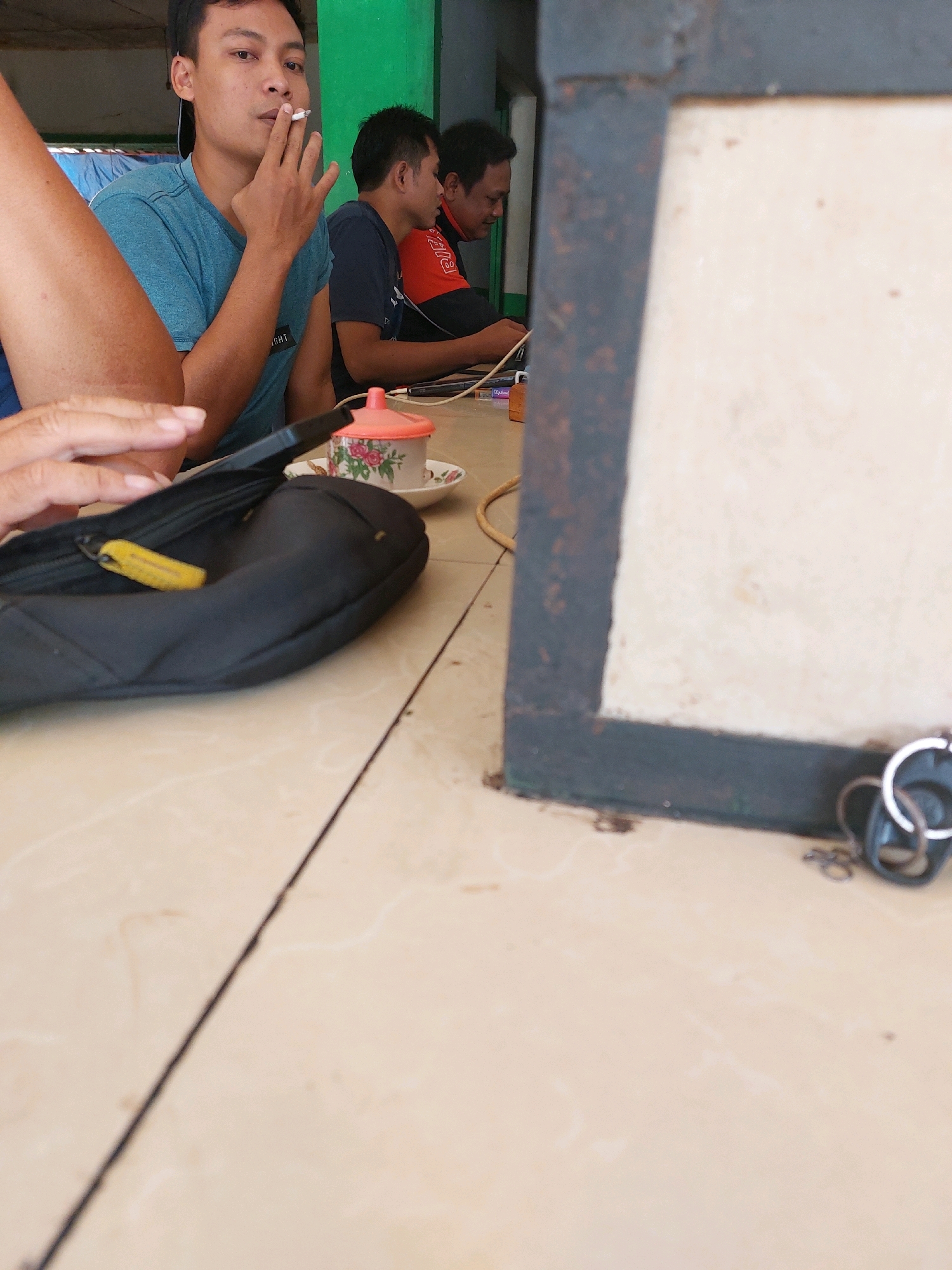 In Sumuragung, Indonesia, a medium shot captures a table scene with four individuals. In the foreground, a person's left arm and hand are visible, holding a dark-colored pouch or bag with a yellow zipper pull. To the left of this, a man in a blue t-shirt is in focus, smoking a cigarette with his right hand raised to his mouth. Behind him, two other men are seated, both looking down at something on the table, with one wearing a dark t-shirt and the other an orange and black jacket. The environment appears to be an outdoor seating area with a light-colored tiled floor and a green wall in the background. A metal frame with a light-colored surface is visible on the right side of the image, and a set of keys hangs from it. A small teacup with a floral pattern is also on the table.