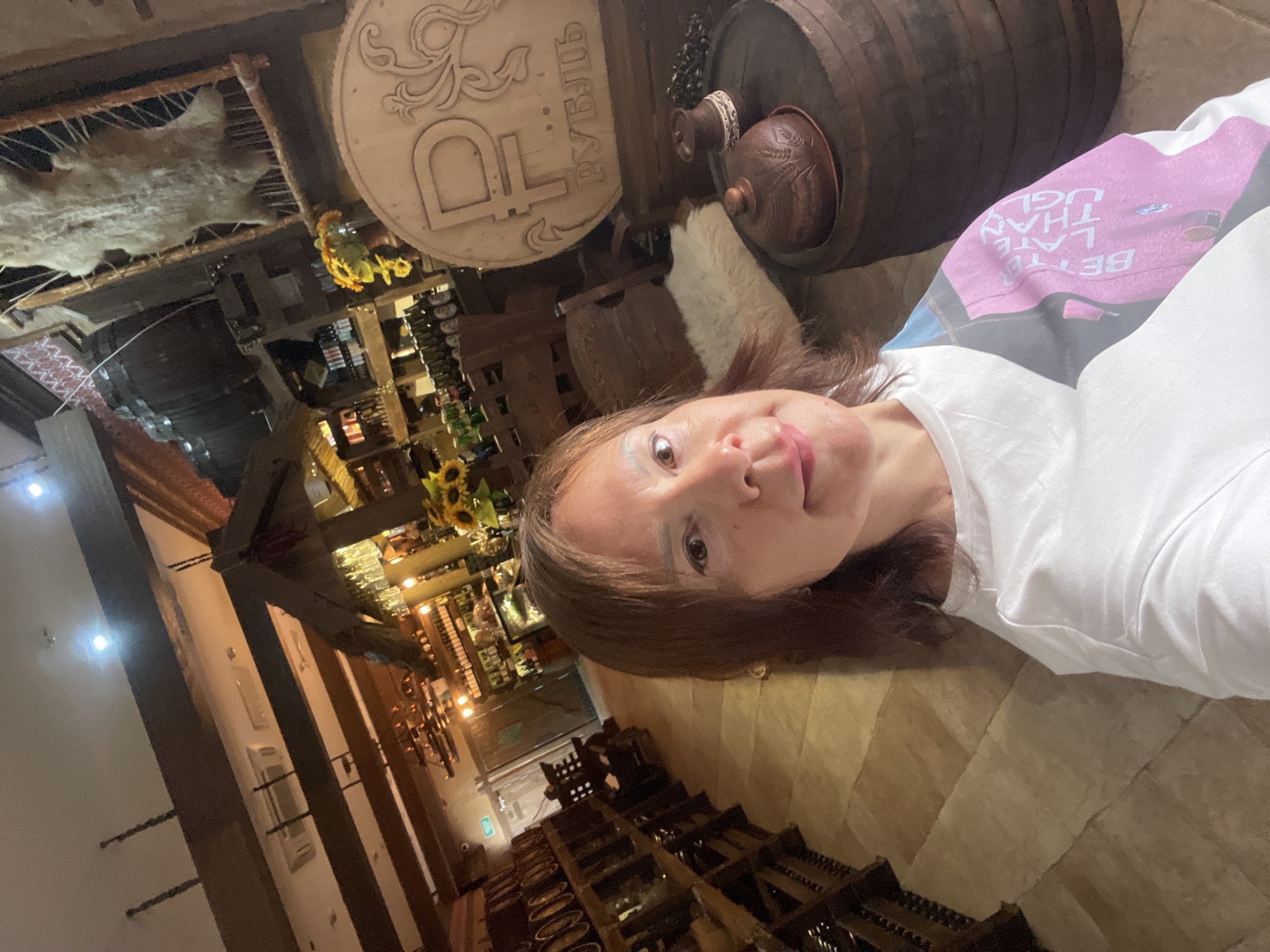 The image is a selfie taken by a woman in a restaurant or bar in Estosadok, Russia. The woman is wearing a white t-shirt with a colorful graphic on the sleeve. She has shoulder-length brown hair and is looking at the camera with a slight smile. The background of the image shows the interior of the establishment, which appears to have a rustic theme. There are wooden beams on the ceiling, shelves stocked with bottles, and decorative elements like a large wooden coin with a ruble symbol, barrels, and what appears to be a hide hanging on the wall. The lighting is warm and atmospheric.