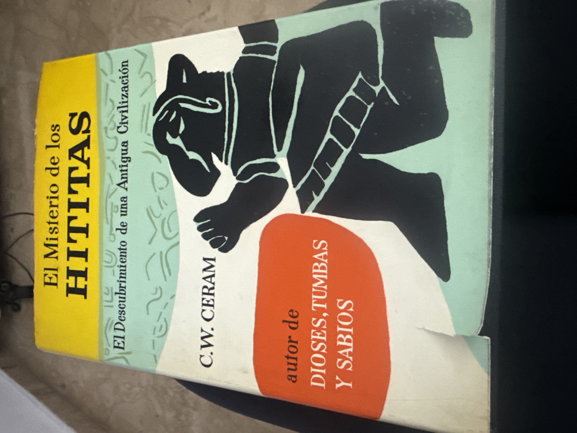 The image shows the side of a book titled "El Misterio de los Hititas" (The Mystery of the Hittites) by C.W. Ceram. The subtitle is "El Descubrimiento de una Antigua Civilización" (The Discovery of an Ancient Civilization). It also states that the author is the author of "Dioses, Tumbas y Sabios" (Gods, Tombs, and Sages).
The spine is yellow, transitioning to a light green, and then to a cream/white color with black line art depicting a stylized figure. There is a tear at the bottom edge.