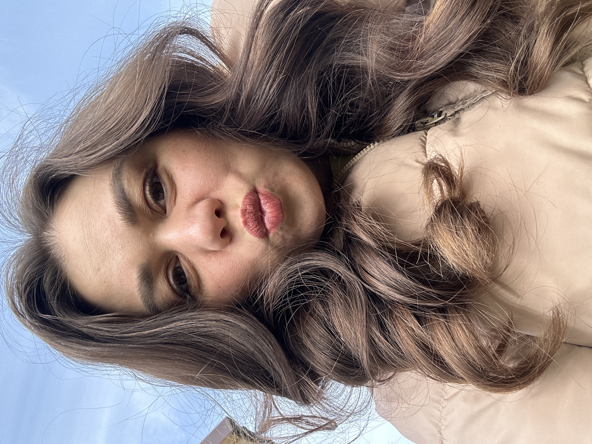 The image shows a young woman captured in a low-angle selfie, with her head tilted diagonally across the frame. She has long, voluminous, dark brown hair with lighter highlights, which cascades around her face and over her shoulders. Her dark eyes are looking directly at the camera, and her lips are pursed in a "kiss" or "duck face" expression, conveying a playful mood. Her skin appears fair with some natural redness around the nose and cheeks, and her eyebrows are well-defined.
She is wearing a light beige or tan puffer-style jacket, which is visible on the right side of the image, with a silver zipper pull visible near her shoulder. The background consists predominantly of a clear, bright blue sky, indicating an outdoor, daytime setting. A small, dark architectural detail, possibly a building or roofline, is subtly visible in the bottom left corner against the sky. The lighting is natural and bright. No visible text is present in the image. The location context is Chaikovsky, Russia.