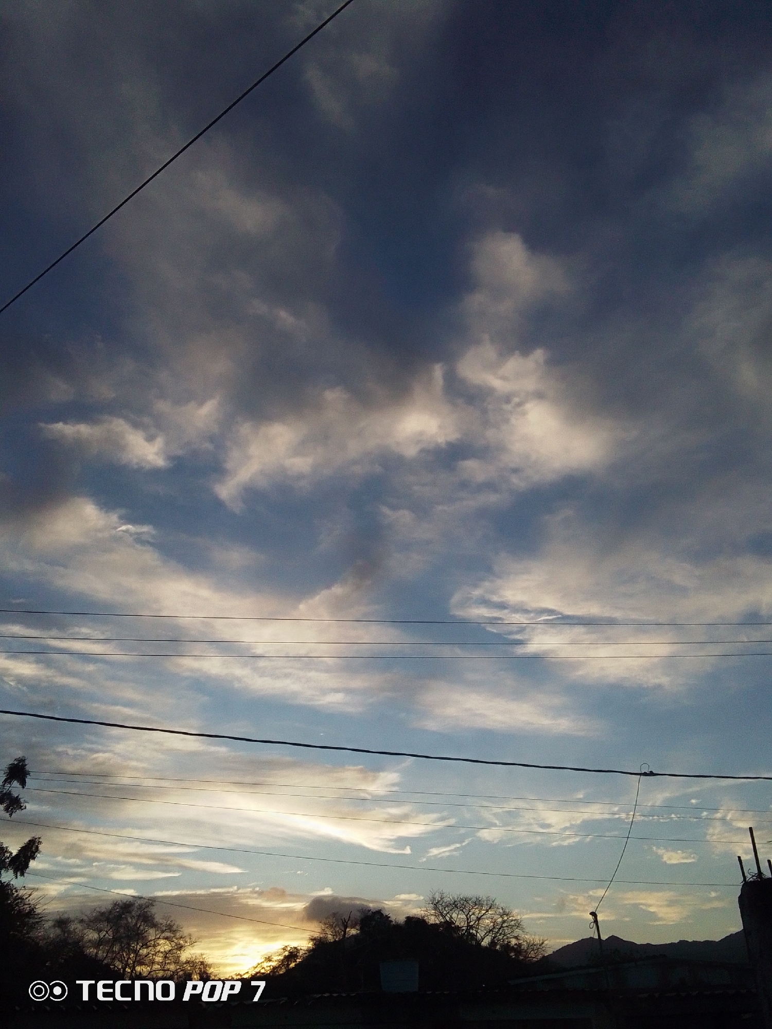 The image shows a dusk or dawn sky with clouds, power lines, and silhouettes of trees and buildings in Altagracia, Venezuela. The sky transitions from darker, overcast clouds at the top to lighter, streaky clouds near the horizon where a faint light suggests the sun. Several power lines traverse the scene horizontally. Silhouetted trees and rooftops occupy the bottom portion of the image, giving a sense of a residential area. The mountains are in the distance.