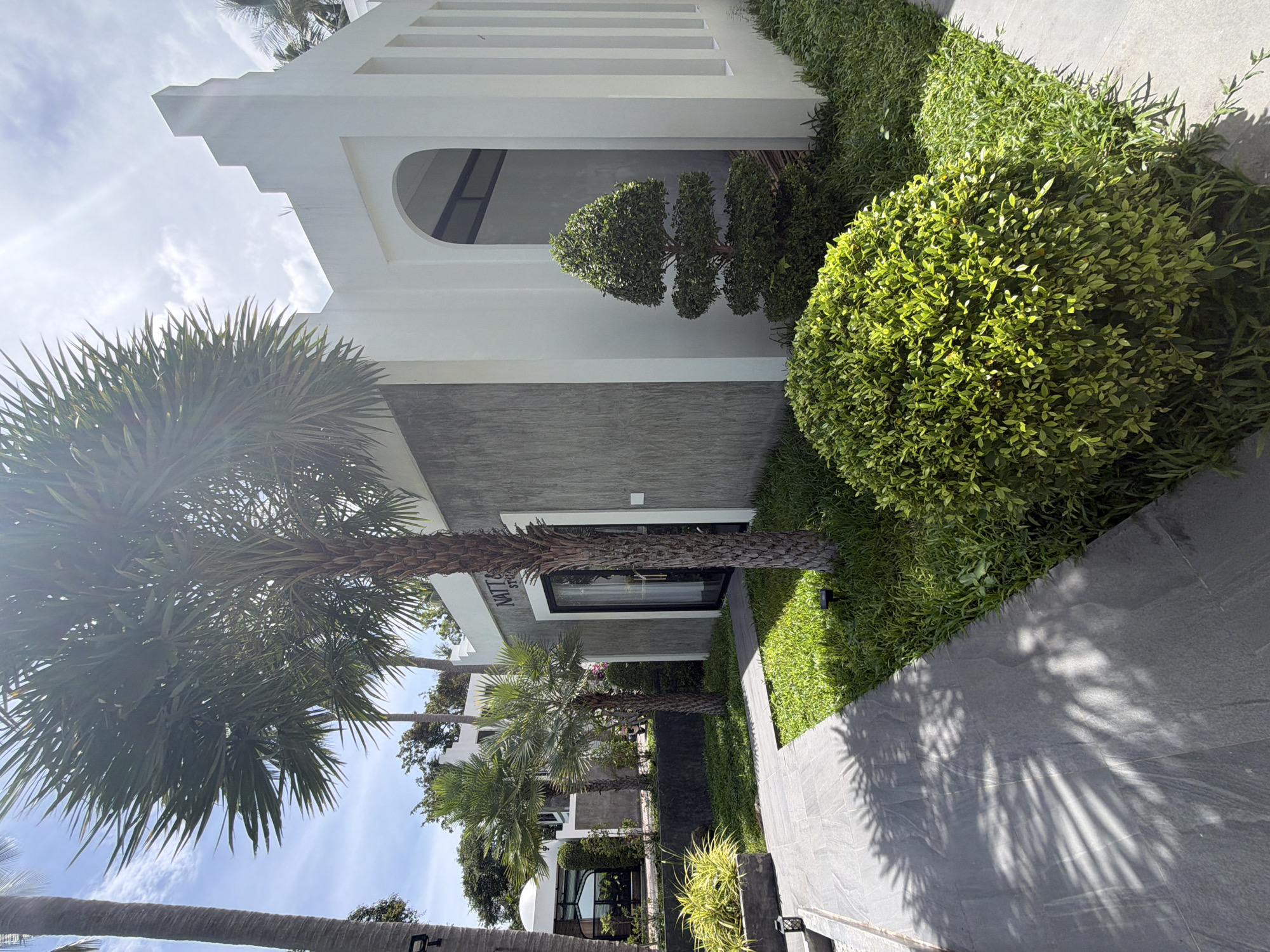 The image shows an outdoor scene with a modern white building and lush greenery. A palm tree trunk is prominently in the foreground, with its fronds extending upwards. To the right of the palm tree is the side of the building, featuring a textured gray wall and a white architectural element with horizontal slats. Further to the right, there is a neatly manicured, spherical green bush and a row of smaller green plants. A paved pathway made of gray stone runs alongside the greenery, with dappled shadows from the palm fronds cast upon it.
The building has an arched opening revealing a dark interior. Above the arched opening, the white structure continues with a series of horizontal white bands. The overall aesthetic suggests a contemporary architectural design. The scene appears to be in a tropical or subtropical location, given the presence of palm trees. The lighting and shadows indicate it is daytime, likely with bright sunlight.
There is visible text on a white strip above a window or doorway on the gray section of the building, which reads "NATT & SOIE". This text, along with the context of Ko Pha Ngan, Thailand, suggests this might be the entrance or signage for a business or establishment. The image evokes a sense of tranquility and well-maintained landscaping.
