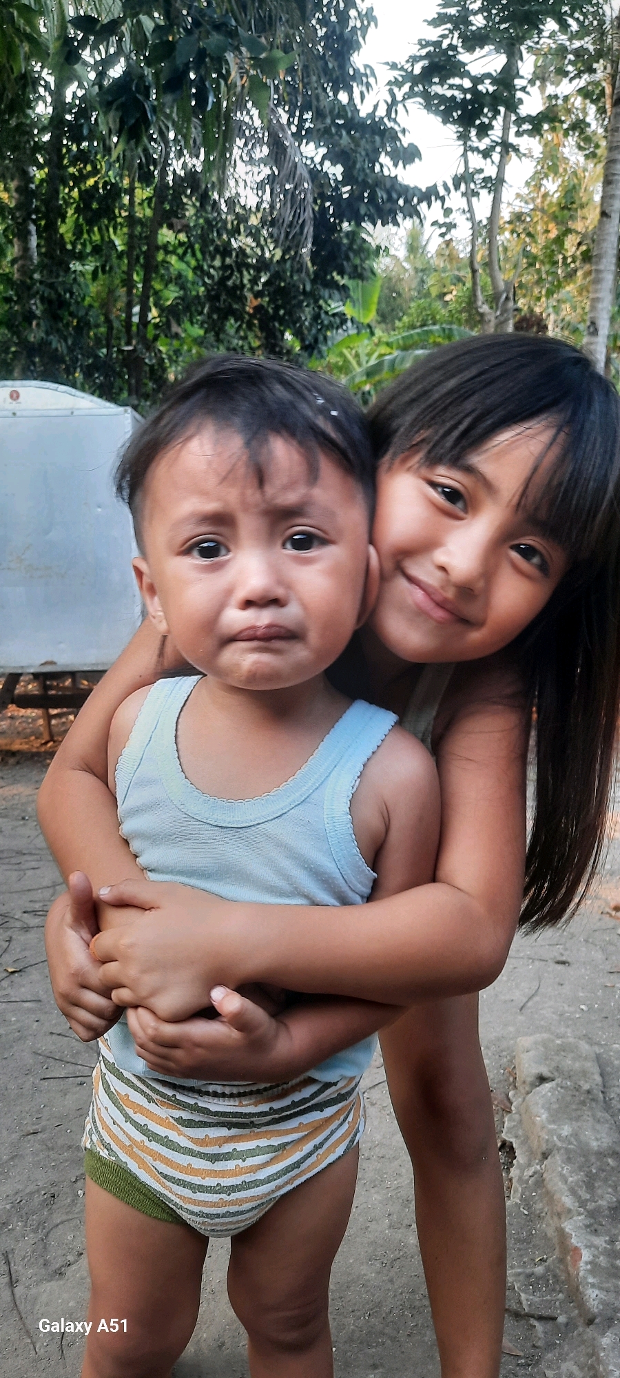 In Jatitengah, Indonesia, two young children are pictured in a close embrace. The younger child, a boy, is in the foreground, wearing a light blue tank top and striped briefs, and appears to be crying with tears visible on his cheeks. The older child, a girl, is behind him, embracing him from behind and smiling warmly. The setting appears to be outdoors, with trees and vegetation in the background, suggesting a rural or semi-rural environment. A white structure, possibly a shed or building, is visible to the left. The ground is dirt or gravel. The overall impression is one of sibling affection, despite the younger child's distress.