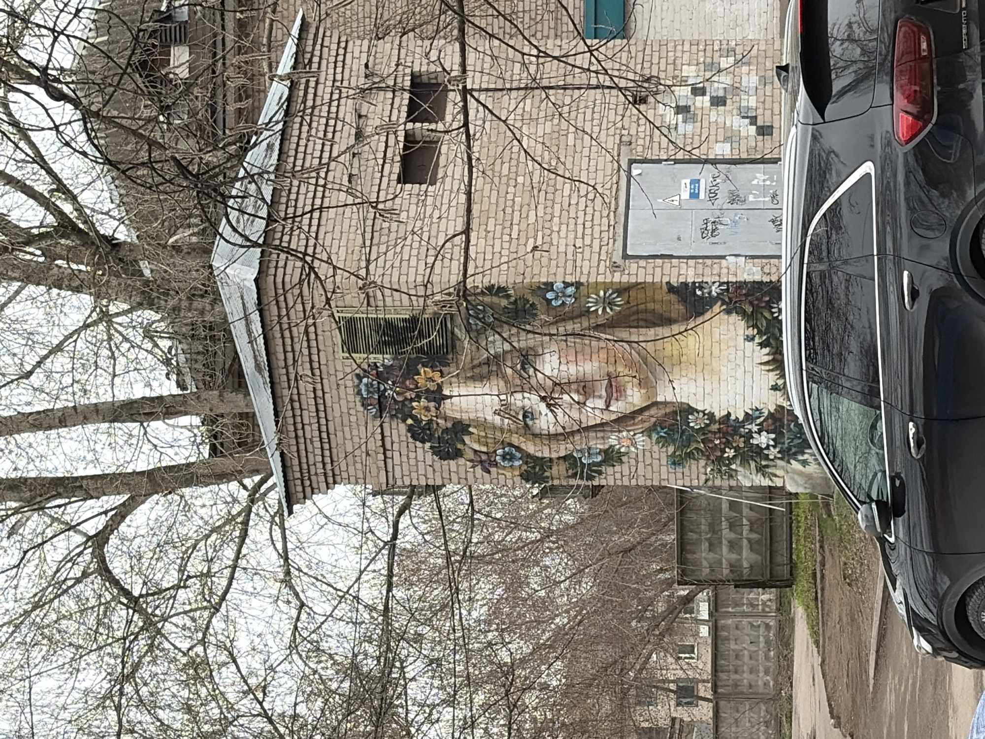 This image presents an urban street scene captured on an overcast day, likely in late winter or early spring, given the leafless deciduous trees. The primary subject is a two-story brick building, which features a large and vibrant mural on its facade. The mural depicts a serene-faced woman, adorned with an elaborate wreath or crown of various flowers and green foliage, suggesting a nature-inspired theme.
To the right, a dark-colored Kia Sorento SUV is parked, with its rear portion, including a distinctive red taillight and the "SORENTO" emblem, clearly visible. The vehicle's reflection shows the surrounding environment. The street below appears to be a mix of pavement and dirt, with small patches of dry grass.
The brick building also has a small, dark rectangular window opening and a metallic grate on its lower section. Above the mural and to the left of the car, a gray utility door or panel is mounted on the brick wall. This panel displays a yellow triangular warning sign with a black lightning bolt, indicating an electrical hazard, and is covered with several instances of indistinct graffiti. Bare tree branches, some with tiny buds, frame the scene from the left and top, silhouetted against a bright, uniform sky. In the background, beyond the car and utility door, concrete block fences delineate property lines. The overall atmosphere is quiet and subdued, with the colorful mural serving as a striking artistic element.