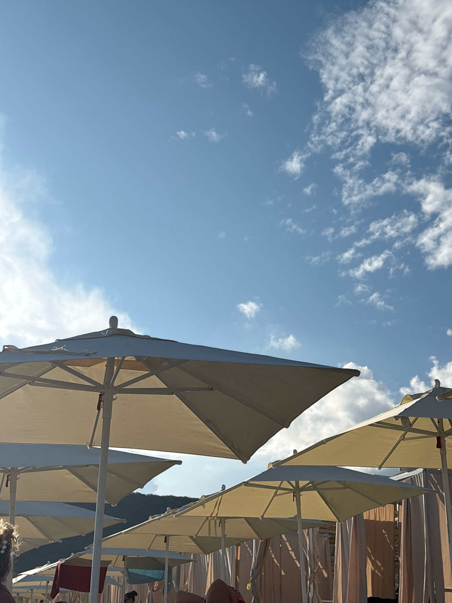 The image captures a sunny day at a beach in Arkhipo-Osipovka, Russia. Several beach umbrellas, some open and some closed, are set up, providing shade. In the background, the outline of a hill covered in vegetation is visible against a bright blue sky dotted with fluffy white clouds. The scene suggests a relaxed atmosphere, likely a vacation or leisure spot.