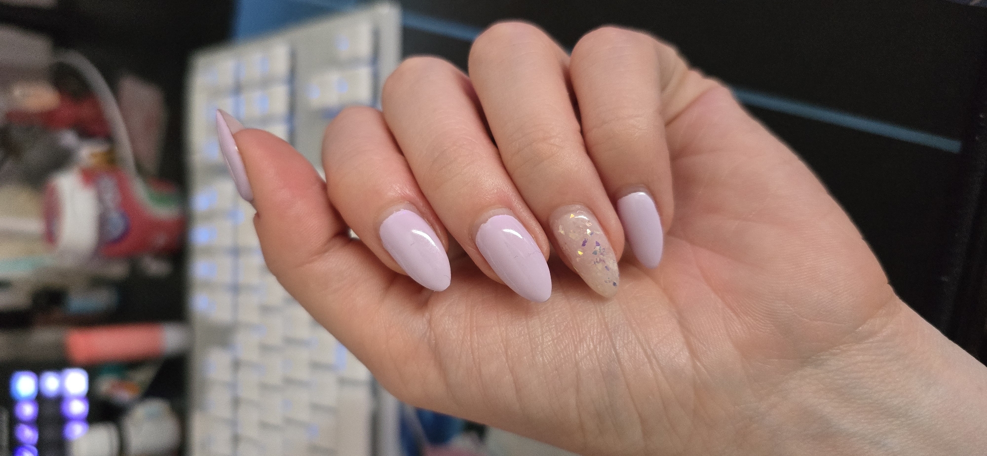 A hand with lavender and glittery nails is displayed in the foreground of the image. The nails are almond-shaped. The background features a blurred view of what seems to be a keyboard and various objects on a desk. The location is not discernible as Burgas, Bulgaria based on the given information.