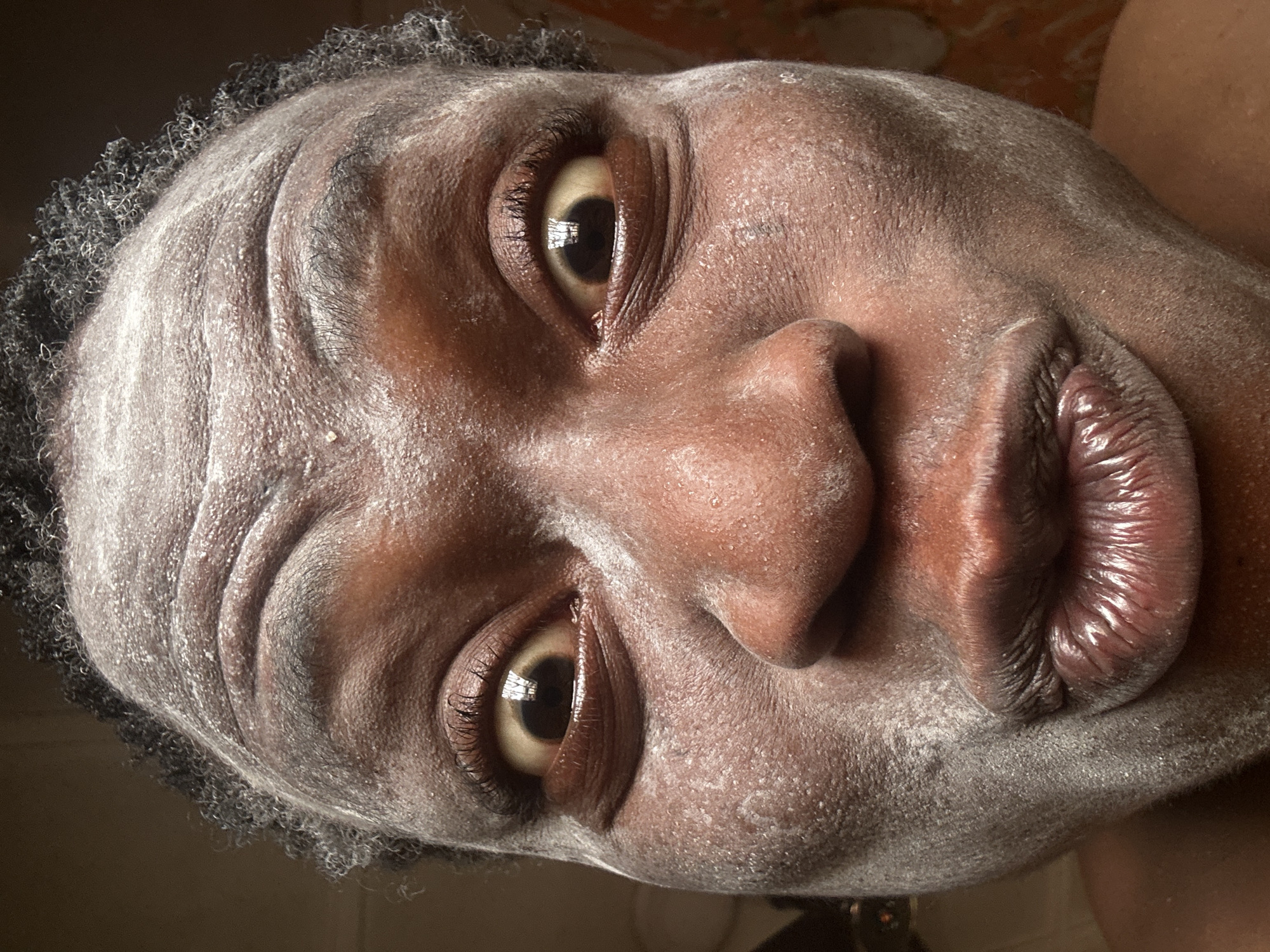 The image shows a close-up of a dark-skinned person's face covered in a white powder. The person is looking directly at the camera with a slight pout. The focus is sharp, highlighting the texture of the skin, the eyelashes, and the details of the eyes and lips. The background is out of focus and provides no location information. No city is identifiable, and the environment is unidentifiable beyond the immediate space around the person's face.