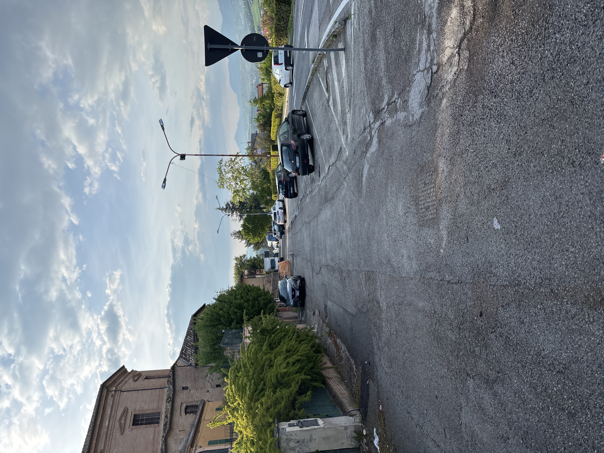 The image captures a street scene in Camerino, Italy, under a partly cloudy sky during daytime. The view looks down a paved asphalt road that shows some cracks and wear, with white road markings visible on the right.
On the right side of the road, several cars are parked in a row. A dark hatchback is prominent in the mid-ground, displaying a partial European license plate "DE 678FJ." Further along, a white hatchback and other vehicles, including vans in the distance, are visible. A black and white triangular "yield" sign is prominently positioned on a pole on the right side of the road, near the parked cars. In the background, distant mountains rise above the horizon.
On the left side of the road, an old, multi-story building constructed of brick and stucco with a red-tiled roof is present. The building features several windows, some with closed green shutters. Lush green bushes and trees line this side of the street, partially obscuring other buildings. A street lamp with two lights extends over the road from the left side, and another lamp is visible further down the street. In the foreground on the left, a concrete post with the house number "8" is visible. No people are present in the scene. The lighting suggests it is daytime, with the sun likely positioned to the left of the frame.