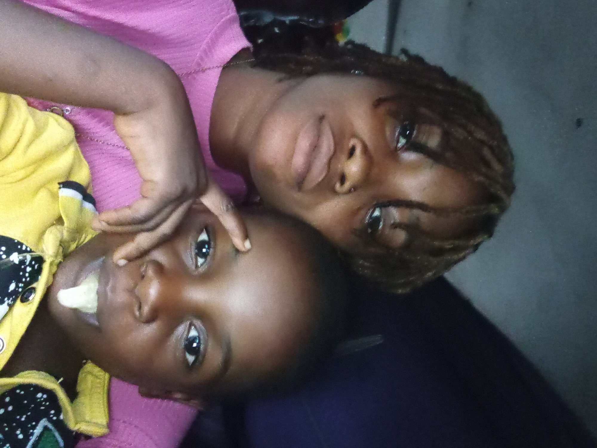 This image captures two individuals, an adult female and a child, in a close-up, diagonal portrait, likely taken indoors in Oba-Ile, Nigeria. Both subjects have dark skin and are looking towards the camera.
The adult, positioned slightly behind and above the child, wears a pink top and has dark hair styled in dreadlocks or braids. Her expression is neutral to thoughtful. A thin chain is visible around her neck. Her arm is gently placed around the child, with one hand resting near the child's eye.
The child, in the foreground, wears a yellow garment with black and white patterned sleeves. A small, light-colored piece of food, possibly a banana or plantain, is held in their mouth. The child has wide, dark eyes and a neutral expression.
The background is dark and indistinct, suggesting an intimate indoor setting with subdued lighting, possibly indicating evening or a room with limited natural light. The overall scene conveys a sense of warmth and closeness between the two individuals. There is no visible text in the image.