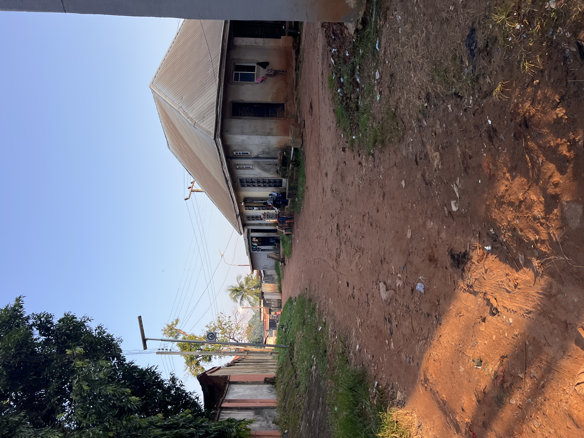 The image depicts a dusty, reddish-brown dirt road in Nkpor, Nigeria. On the right side of the road is a light-colored, single-story building with a corrugated metal roof. The building has several windows and at least two doorways. Clothes are hanging on a line outside one of the windows. In front of the building, there are several chairs and what appears to be a small gathering of people.
To the left of the road, a concrete wall, painted in sections of white and reddish-pink, runs along the side. Beyond this wall, a dense green tree is visible, its branches extending towards the top left of the frame. A street lamp with a solar panel is positioned near the wall. Further down the road, more buildings and palm trees can be seen. The ground on both sides of the road is covered with patches of dry grass and scattered debris, including plastic waste. The sky is clear and blue, indicating daylight, possibly morning or late afternoon, given the angle of the light casting shadows.