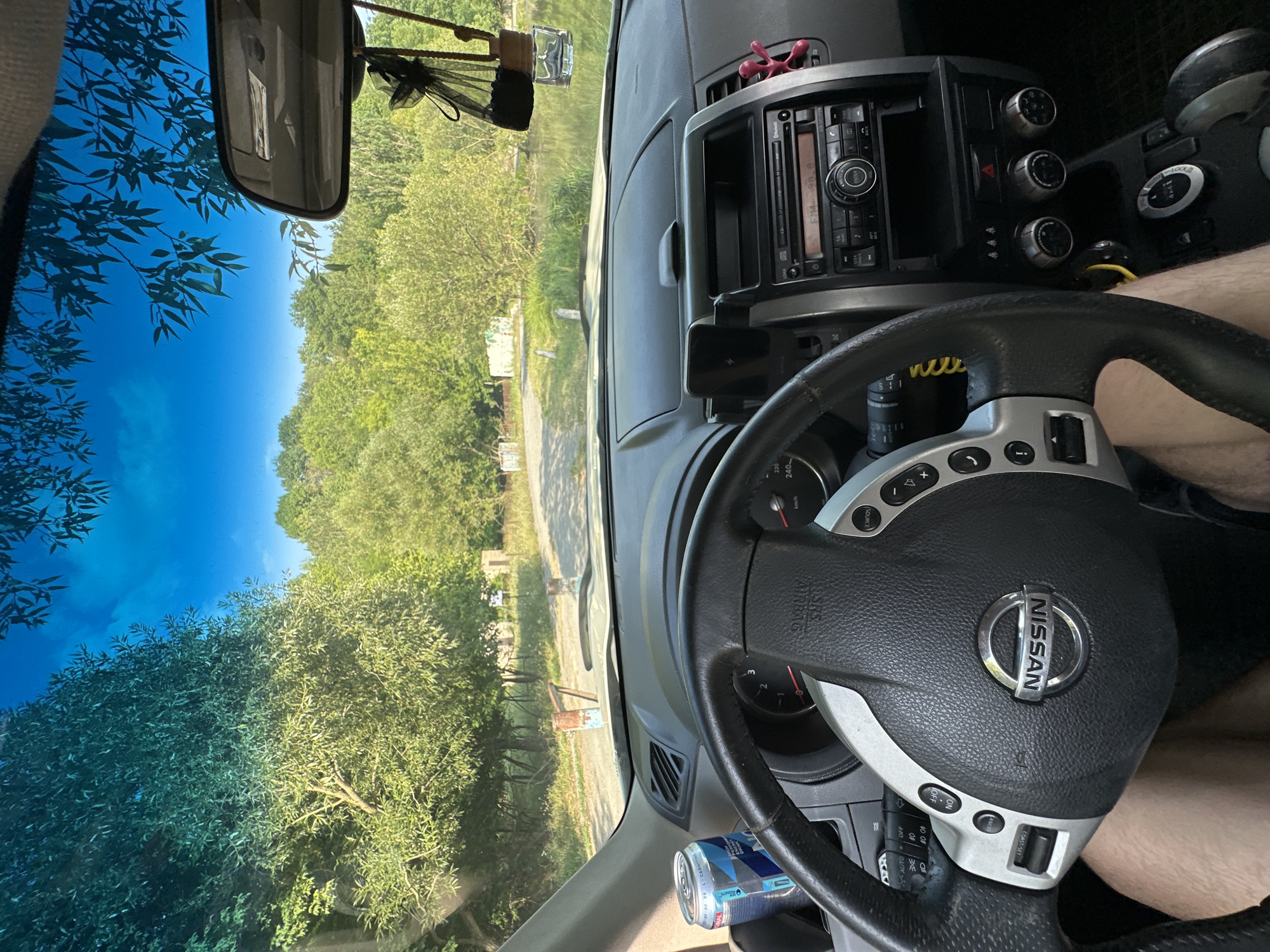 The image is taken from the interior of a Nissan X-Trail, looking forward through the windshield. The driver's side of the steering wheel and dashboard are visible. A can of energy drink is in the cup holder to the left of the steering wheel. Outside, a dirt road leads through a wooded area with a small body of water on the right. The sky is blue with some clouds and tree branches are visible in the foreground, obscuring parts of the sky. The scene appears to be in a rural area, possibly near Makiv, Ukraine. The car is stationary.