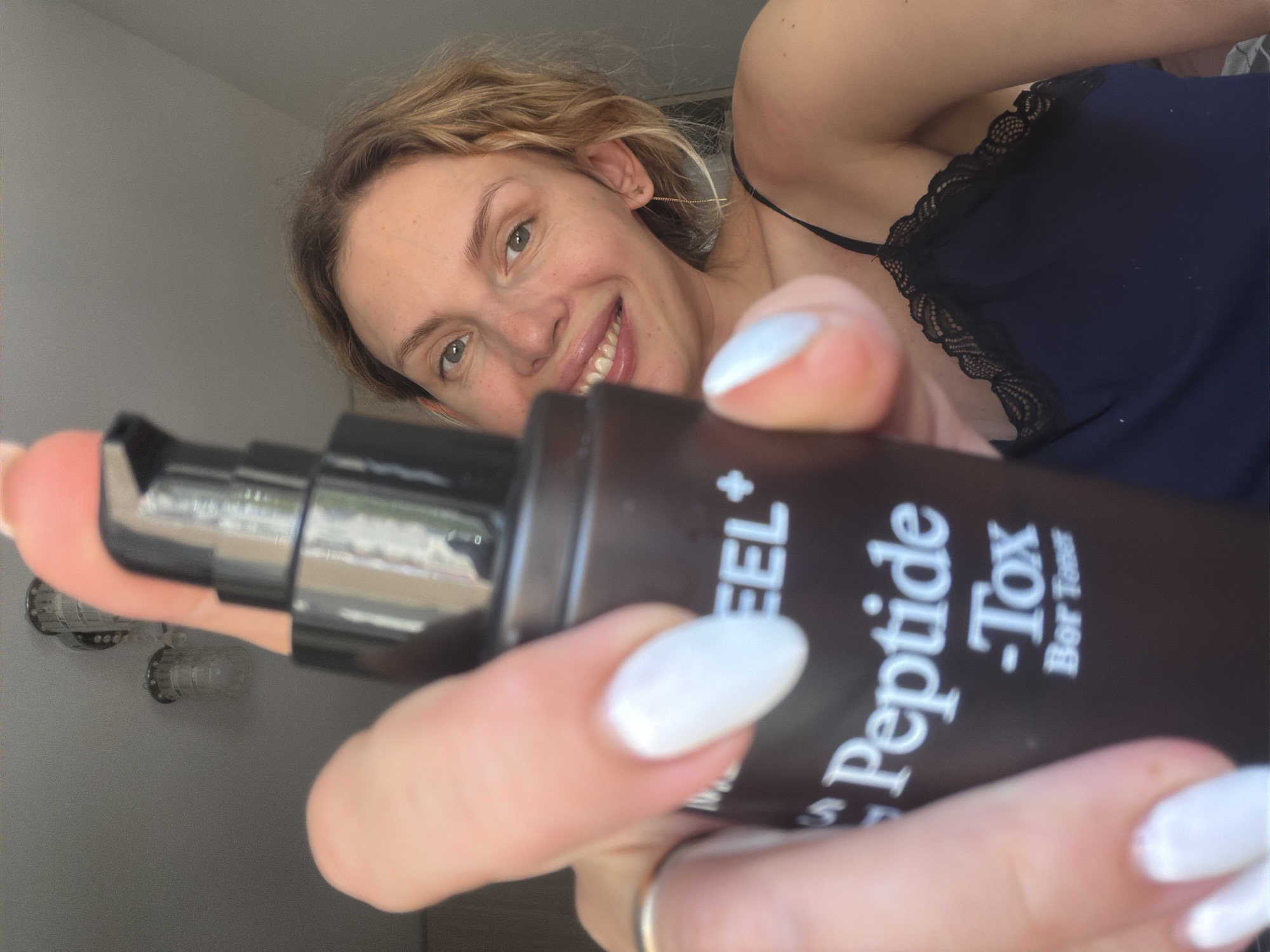 A woman is holding a dark brown bottle labeled "Peptide - Tox Bor Toner". She is smiling and has light skin, blonde hair, and is wearing a dark blue top with lace trim. Her fingernails are painted white. The background is out of focus, but the reflection in the bottle shows a window and what appears to be a light fixture. Based on the reflection and general appearance, it is possible this image was taken indoors. It is not possible to confirm the location is Budva, Montenegro from the image alone.