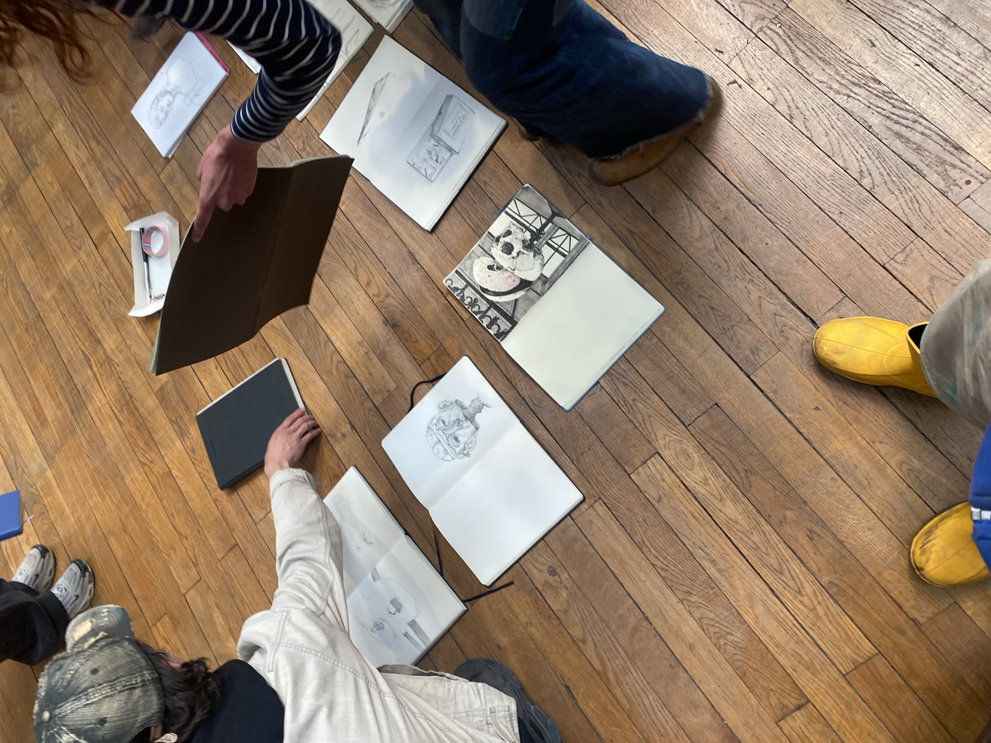 An overhead shot captures several individuals gathered on a light brown wooden floor, engrossed in reviewing various open sketchbooks and art materials. The setting appears to be an informal art critique or sharing session, likely in a studio or communal space in Paris, France.
On the top left, a person wearing a striped long-sleeve shirt has one hand holding open a dark brown sketchbook, while the other hand points towards its contents. Nearby, a small white container holds a pen and a roll of tape. Adjacent to this, a person in blue jeans and light brown moccasin slippers is partially visible.
The wooden floor is covered with multiple open sketchbooks, each displaying different pencil or mixed-media drawings. One sketchbook reveals a detailed drawing of a person's head or bust. Another features a drawing of a package or box, with faint, largely illegible text on it. A more prominent drawing depicts a stylized skull or a black-and-white panda face with graphic elements. Further down, two other sketchbooks showcase drawings of a grotesque or cartoonish head and a more traditional skull. A closed black sketchbook also rests on the floor, and a blue cover (possibly another sketchbook) is visible on the far left.
On the bottom right, a person wearing distinctive yellow rain boots and light-colored pants is seen. To the bottom left, another individual in a dark t-shirt, a light jacket, and a distressed baseball cap is visible, with their hand resting on a black sketchbook. Grey sneakers are seen next to them. The atmosphere suggests a focused and collaborative artistic environment, possibly during an art class or workshop. No specific time of day or weather is discernible.