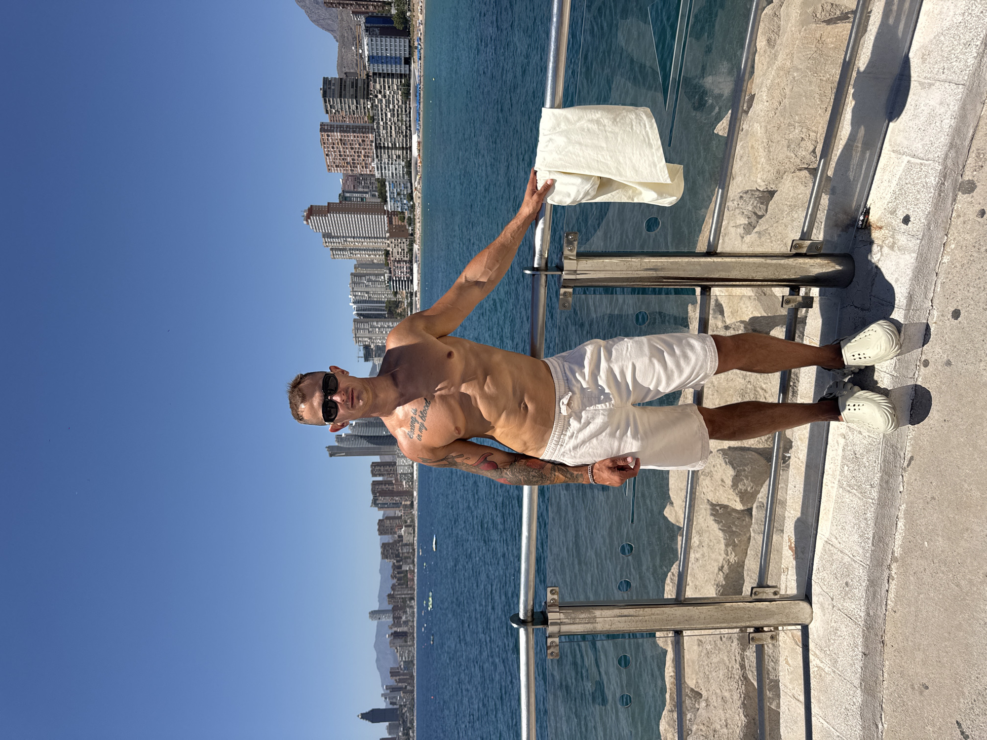 A shirtless man stands on a stone walkway by the sea in Benidorm, Spain, with the city skyline in the background. He is wearing white shorts and light-colored slip-on shoes. The man has sunglasses on and tattoos on his left arm and torso. His left hand is resting on a metal railing, and a white towel is draped over the railing next to him. The sea is blue and calm, with some boats visible on the water. The city skyline features numerous tall buildings and some mountains in the distance. The sky is clear and blue.