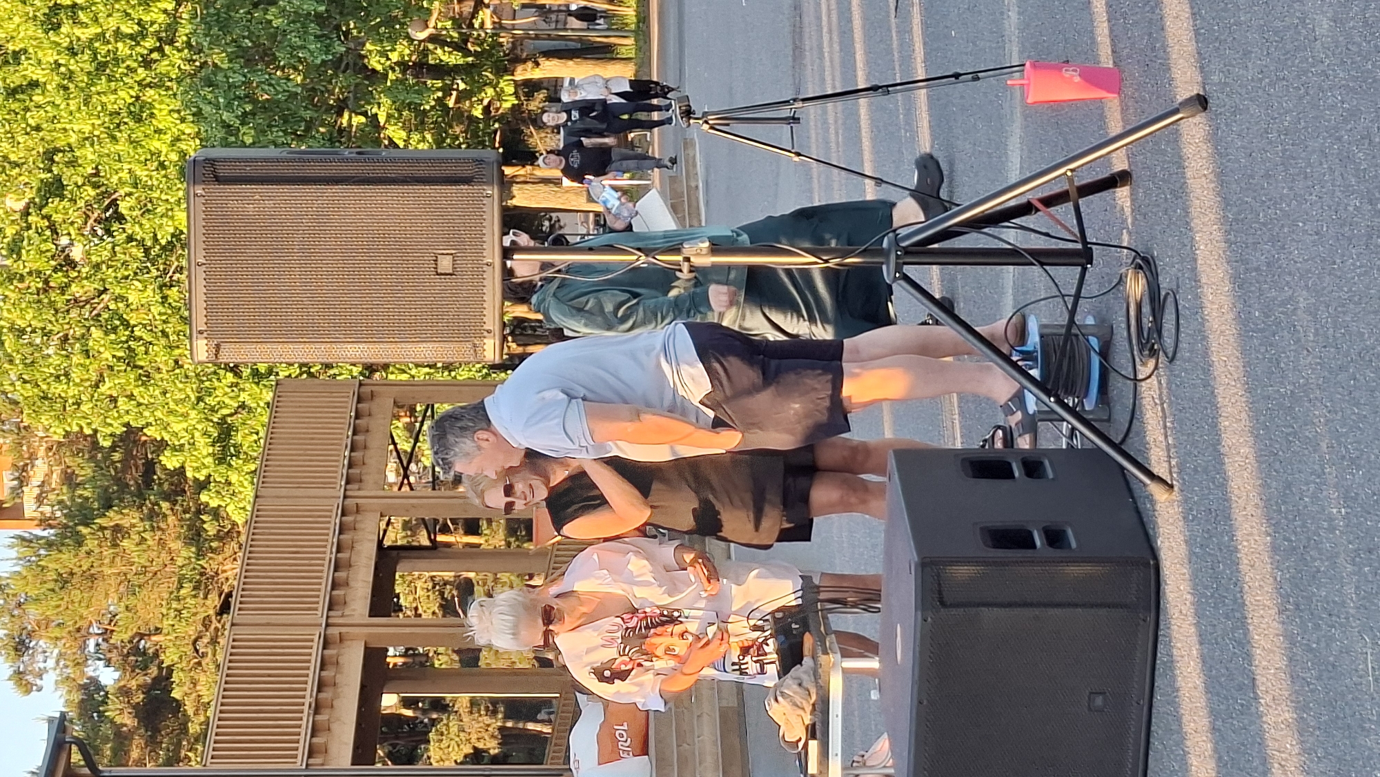 The image captures an outdoor scene in Tallinn, Estonia, likely during the daytime given the sunlight and shadows. In the foreground, a large black speaker is positioned on the ground, with a tripod holding another speaker standing to its right. Various cables and a cable reel are visible around the speaker equipment.
Three people are the central focus in the mid-ground. A woman with blonde hair pulled up in a bun and wearing sunglasses is standing to the left, dressed in a white shirt with a graphic print and dark shorts. She appears to be holding something in her hands. Next to her, a woman with blonde hair, sunglasses, and a black dress is standing and looking towards the man beside her. This man, wearing a light blue polo shirt and dark shorts, is facing the woman in the black dress, engaged in conversation. Behind them, another person in a dark green hooded outfit is standing with a bottle in hand.
In the background, a wooden structure with railings is visible, suggesting a park or public area. Trees with vibrant green foliage line the background, and the pavement indicates an urban setting. Further in the background, other individuals are seen walking, and a tall building is partially visible through the trees. The lighting suggests it might be late afternoon or early evening, casting long shadows across the ground.