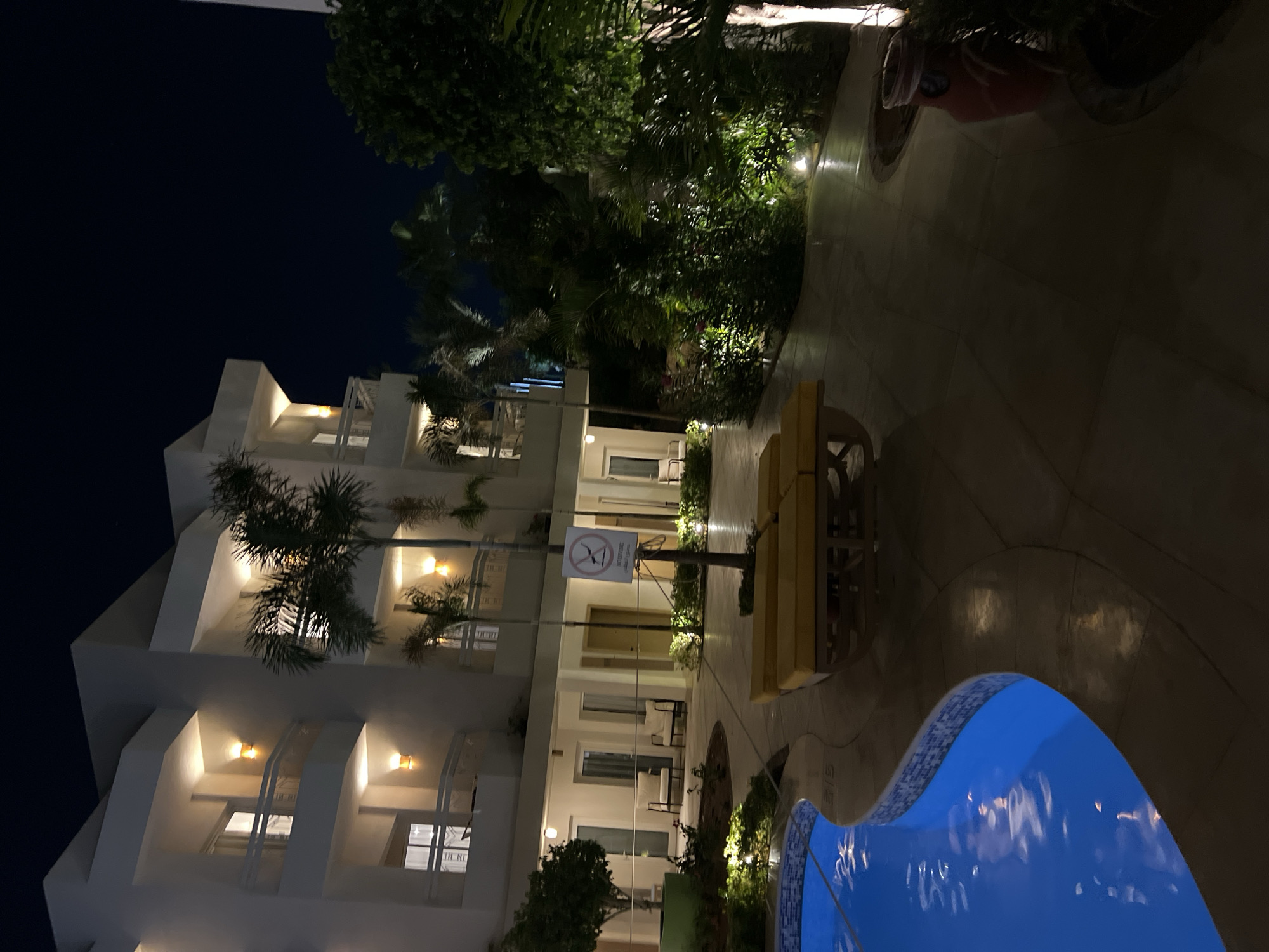 An exterior nighttime shot captures a hotel courtyard in Sharm El Sheikh, Egypt. In the foreground, a bright blue swimming pool curves into the frame on the left, its surface reflecting subtle lights. Adjacent to the pool, two yellow lounge chairs are neatly arranged on a paved patio. A lamppost with a sign prohibiting sitting on the pool deck stands between the lounge chairs and the hotel building. The hotel itself is a multi-story white structure with balconies and windows illuminated by warm light. Tropical palm trees and lush green foliage surround the courtyard, with accent lighting highlighting the plants. The overall scene suggests a tranquil resort setting at night.