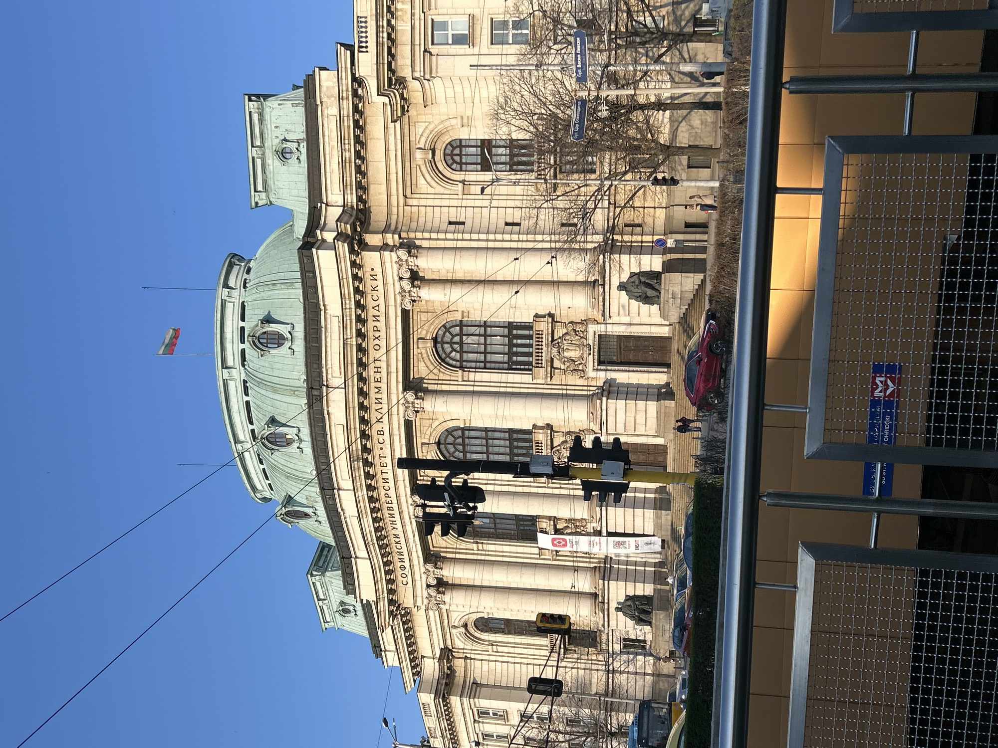 The image shows the facade of Sofia University "St. Kliment Ohridski" in Sofia, Bulgaria, taken from street level. The building is large and ornate, with a prominent dome on top and multiple windows and columns. Red Bulgarian flag is waving from the top of the dome. In front of the building, there are traffic lights, various street signs, and a statue. Several parked vehicles are visible. A metal barrier is in the foreground, partially obstructing the view. People can be seen walking on the steps in front of the building. The sky is clear and blue.