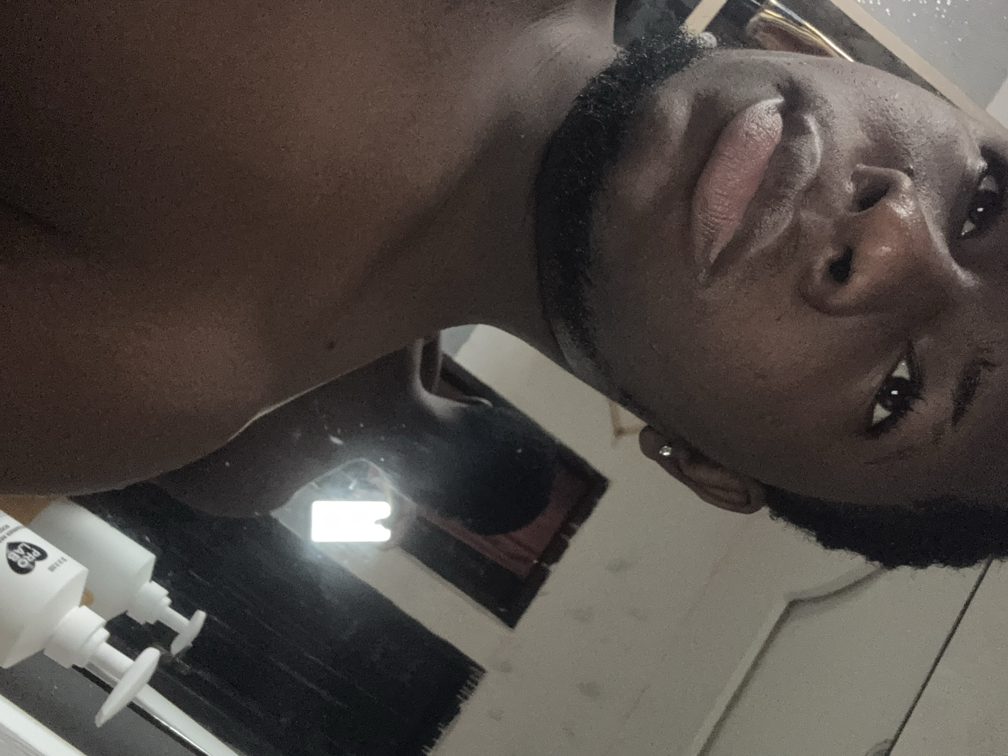 The image captures a mirror selfie of an individual with dark skin, positioned from a low, upward-angled perspective. The person's face is centered, looking directly into the camera with a neutral expression, featuring full lips that are slightly parted, and their visible right eye. Dark facial hair forms a beard or stubble around their jawline, and a small stud earring is visible in their right ear. The upper part of their chest and shoulder are bare.
The setting is an indoor space, likely a bathroom or bedroom, evident from the prominent mirror reflection. In the lower part of the reflection, two white pump dispenser bottles are visible on a surface, possibly a counter or shelf. The text "PRO LAB" is clearly discernible on one of these bottles. Further in the background of the reflection, a dark, rectangular object (perhaps a frame or window) reflects a bright light source. The overall lighting is somewhat dim, creating subtle highlights on the person's skin. The original image is notably tilted. This scene takes place in Ipetumodu, Nigeria.