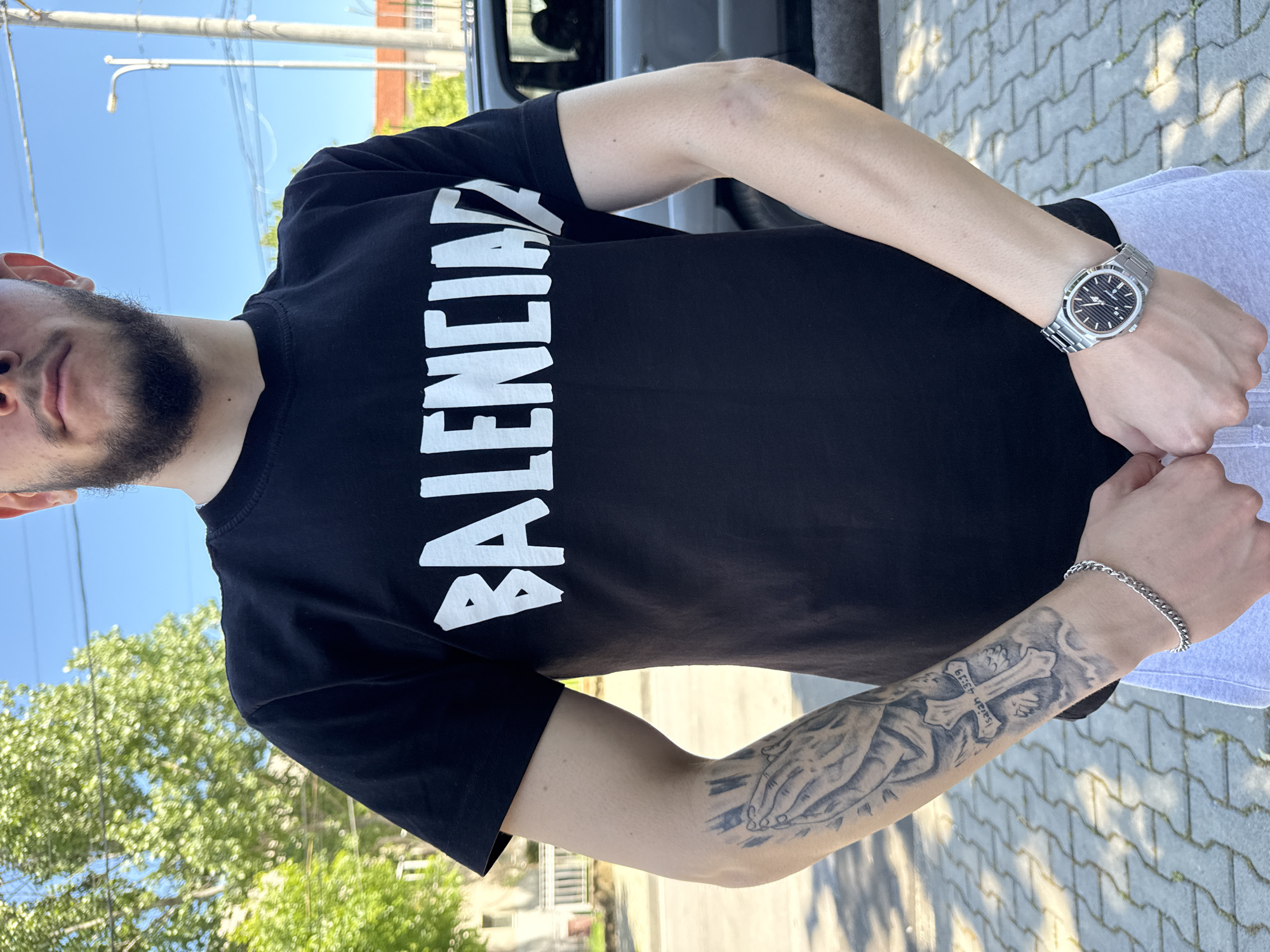 A person wearing a black T-shirt with "BALENCIAGA" printed in white is standing on a gray brick surface. The person has a watch on their left wrist and a tattoo on their left forearm. The background includes a green tree, a blue sky, and utility poles with wires. The location is likely outside. The city is Elhovo, Bulgaria.