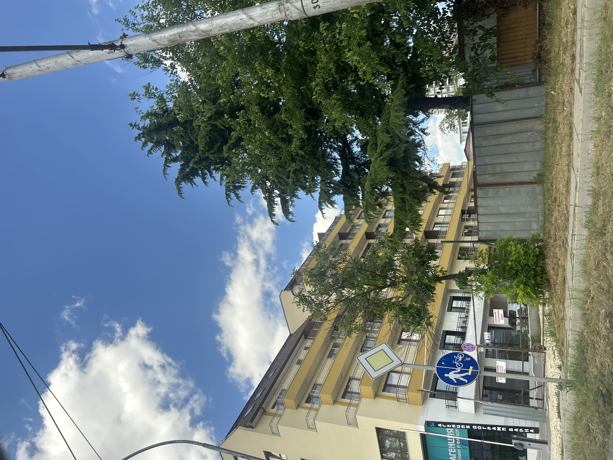 The image depicts a street view in Varna, Bulgaria, on a sunny day with scattered white clouds. In the foreground, a grassy verge borders a paved road. To the left, a modern, multi-story building with a light yellow facade and yellow balconies is visible, alongside a commercial storefront with glass doors and signage in Bulgarian. A vertical barrier, possibly a gate or a retractable bollard, is positioned at the entrance of the storefront. A tall metal pole with wires is present on the right side of the image.
In front of the building, a traffic sign with a yellow diamond shape on a white background is prominently displayed on a pole. Below it, a circular blue traffic sign indicates a shared pedestrian and bicycle path, with a white silhouette of a person walking and a bicycle. Another circular sign with a red border and a red horizontal bar signifies "no entry."
A tall, mature evergreen tree with dense foliage stands in the middle ground, partially obscuring the building. Another smaller deciduous tree with green leaves is also present. Behind the evergreen tree, a grey metal fence runs along the property line, with a section of a brown wooden fence visible to its right. The fence encloses an area where construction or renovation may be taking place, as indicated by some materials leaning against it and a white sign on a gate that reads "ВНИМАНИЕ! СТРОИТЕЛЕН ОБЕКТ" (ATTENTION! CONSTRUCTION SITE). In the background, other buildings and more trees are visible under the bright blue sky. The overall scene suggests an urban environment with a mix of residential, commercial, and potentially developing spaces.