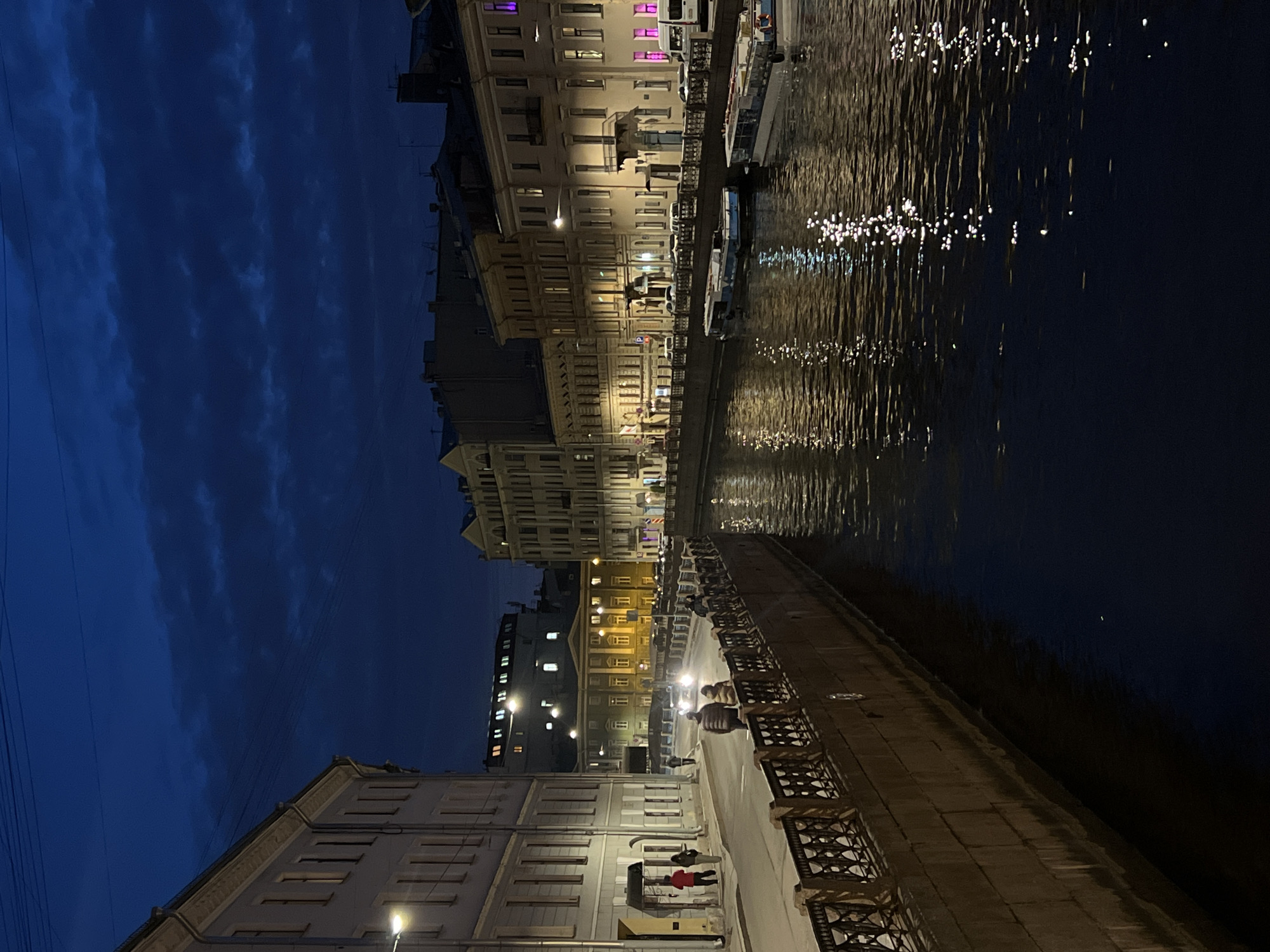 The image depicts a nighttime scene in Saint Petersburg, Russia. A canal runs along the right side of the frame, reflecting the lights from the buildings on the opposite bank, creating shimmering patterns on the water's surface. Buildings line the canal, illuminated with warm light from windows and streetlights. A walkway runs along the near side of the canal with chairs, people can be seen walking, sitting, and interacting on the walkway. The sky above is a deep blue, with light clouds. Power lines are visible in the top left corner. Overall, the image captures a tranquil night along one of Saint Petersburg's canals.