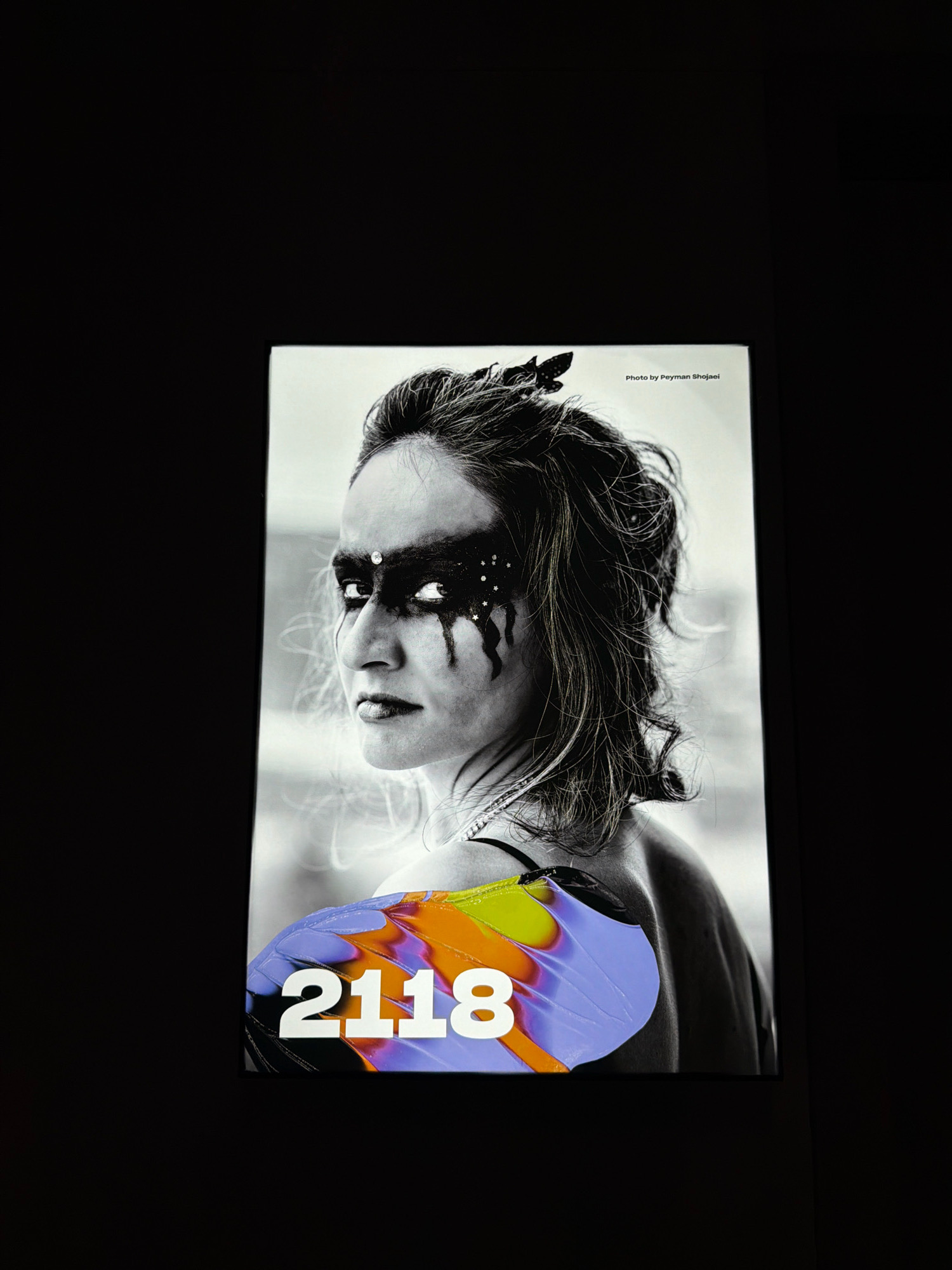 The image shows a close-up of a person's face and shoulder, overlaid with a vibrant, colorful butterfly wing graphic and the number "2118". The person has dramatic dark eye makeup with white accents resembling stars and dripping paint. The background is a stark black. There is no discernible location in the city of Amsterdam, Netherlands.
