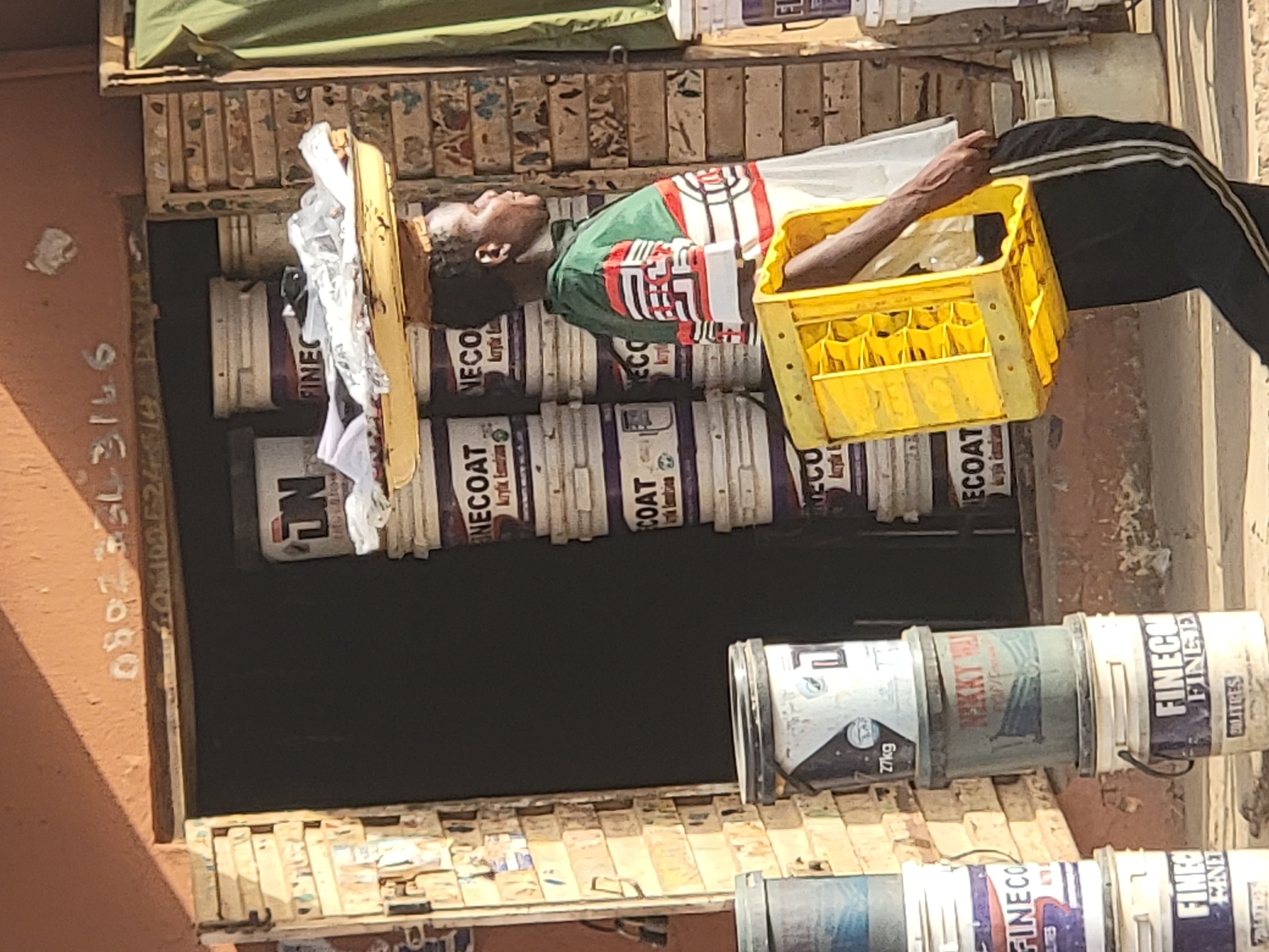 A man is lying on top of a stack of paint cans in front of a shop. He is wearing a green, white, and red shirt with black pants. He is holding a yellow crate in his right hand. The shop appears to be selling paint, as evidenced by the stacks of paint cans. There are phone numbers painted on the building in the left of the image. The location is identified as the city of Maiburiji, Nigeria.