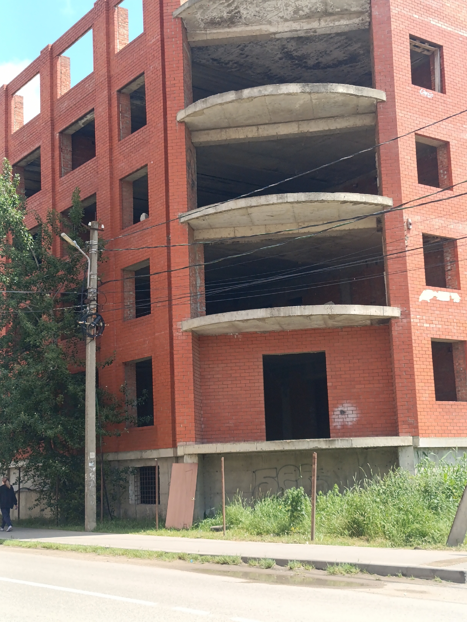 The image shows an unfinished, multi-story brick building with open window spaces in Yablonovskiy, Russia. Concrete balconies are visible on one section of the building. A street with a sidewalk runs along the front of the building. Vegetation grows along the base of the building. A utility pole stands near a tree on the left side of the frame. A person is walking on the sidewalk. A door leans against the building's foundation. Graffiti marks the concrete foundation.