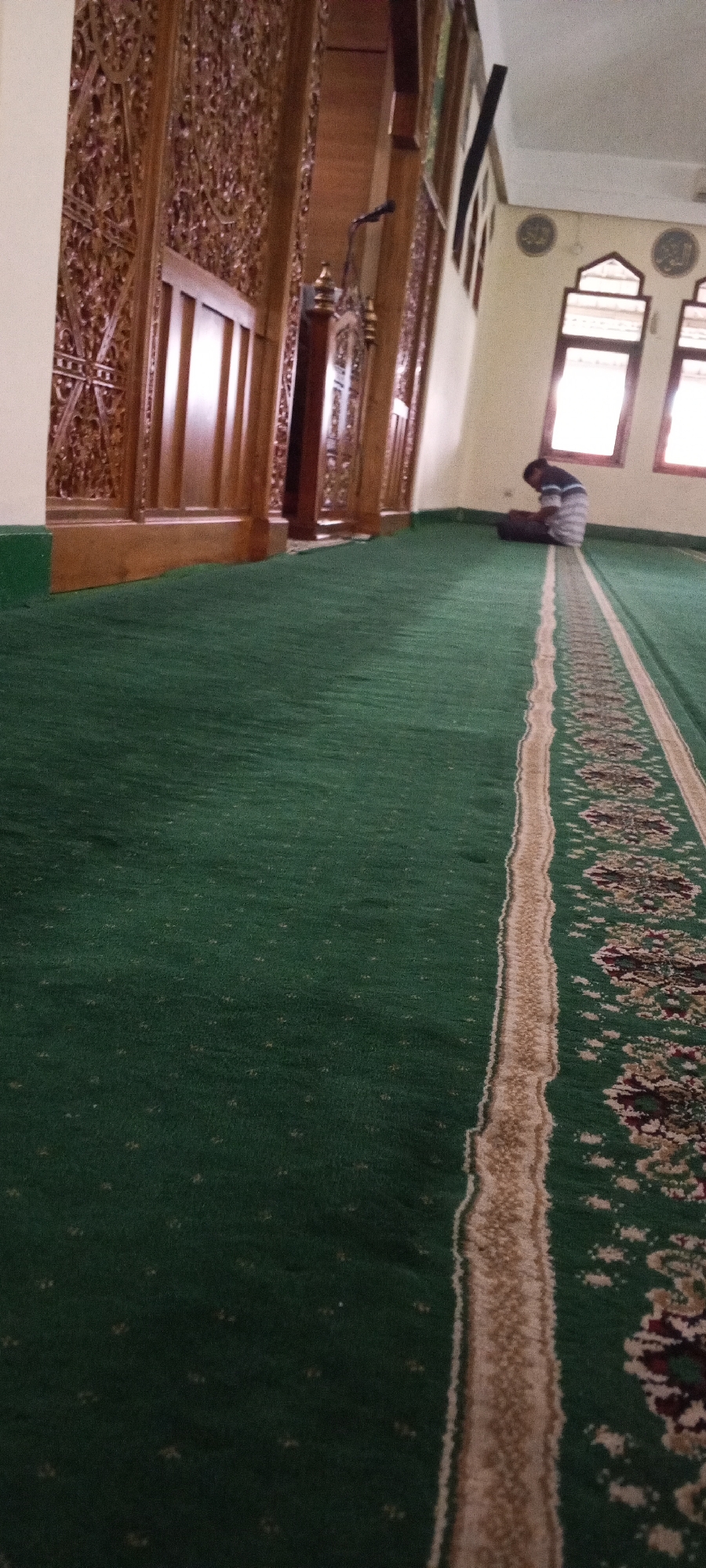 The image depicts the interior of a mosque in South Jakarta, Indonesia. A person is seen performing Islamic prayer, prostrating on a prayer mat placed on a green carpet. The mosque features ornate wooden paneling on the walls, a pulpit, and arched windows allowing natural light. The carpet has a decorative border with intricate patterns.