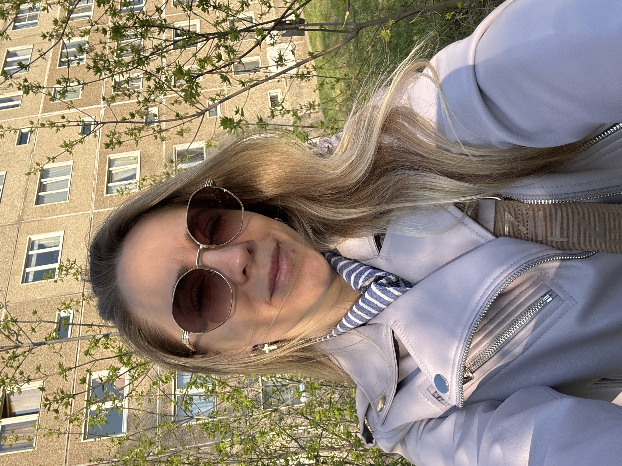 A woman with long, blonde hair is wearing round sunglasses, a light-colored jacket, and a blue and white striped scarf. She is standing outdoors with a multi-story building behind her, possibly an apartment complex, with several visible windows. Trees with budding green leaves are in the foreground, partially obscuring the building. The photo appears to be taken in the city of Chaikovsky, Russia.