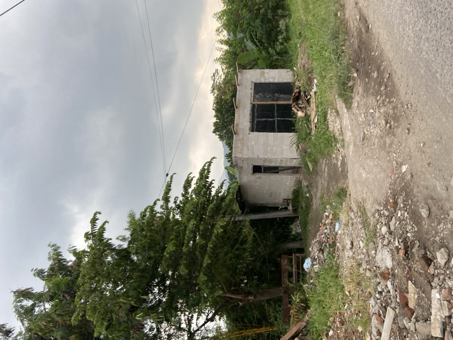 The image shows a small, unfinished concrete building in the middle of a dirt path. The path is located in the city of Kecamatan Margaasih, Indonesia, at coordinates -6.964 latitude and 107.539 longitude. The building has a single door and a window covered with wooden planks. There are several trees and plants in the background, including a large tree on the left side of the image. The ground is covered in dirt, rocks, and debris. The sky is overcast and gray. There are power lines running across the top of the image. There are no people in the image. There is a wooden bench in the image.