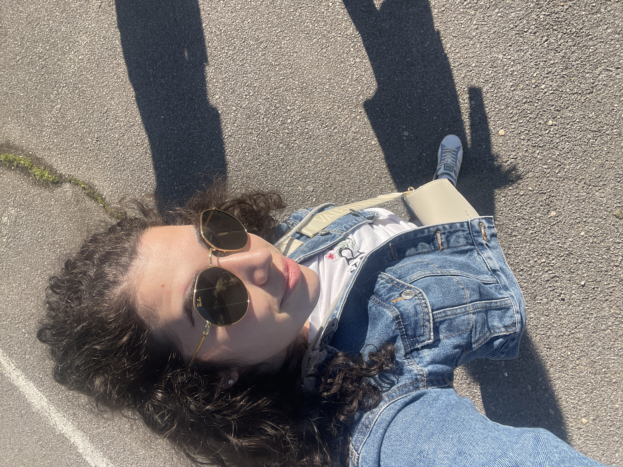 A young woman with curly brown hair and sunglasses is lying on a paved surface, likely asphalt, outdoors. She is wearing a light blue denim jacket and a white t-shirt. A beige purse with a white strap is visible beside her. Her left foot, clad in a grey and white sneaker, is also visible. The scene is bathed in sunlight, casting long shadows from a person or object outside the frame. The location could be a street or parking lot in the city of Sofia, Bulgaria.